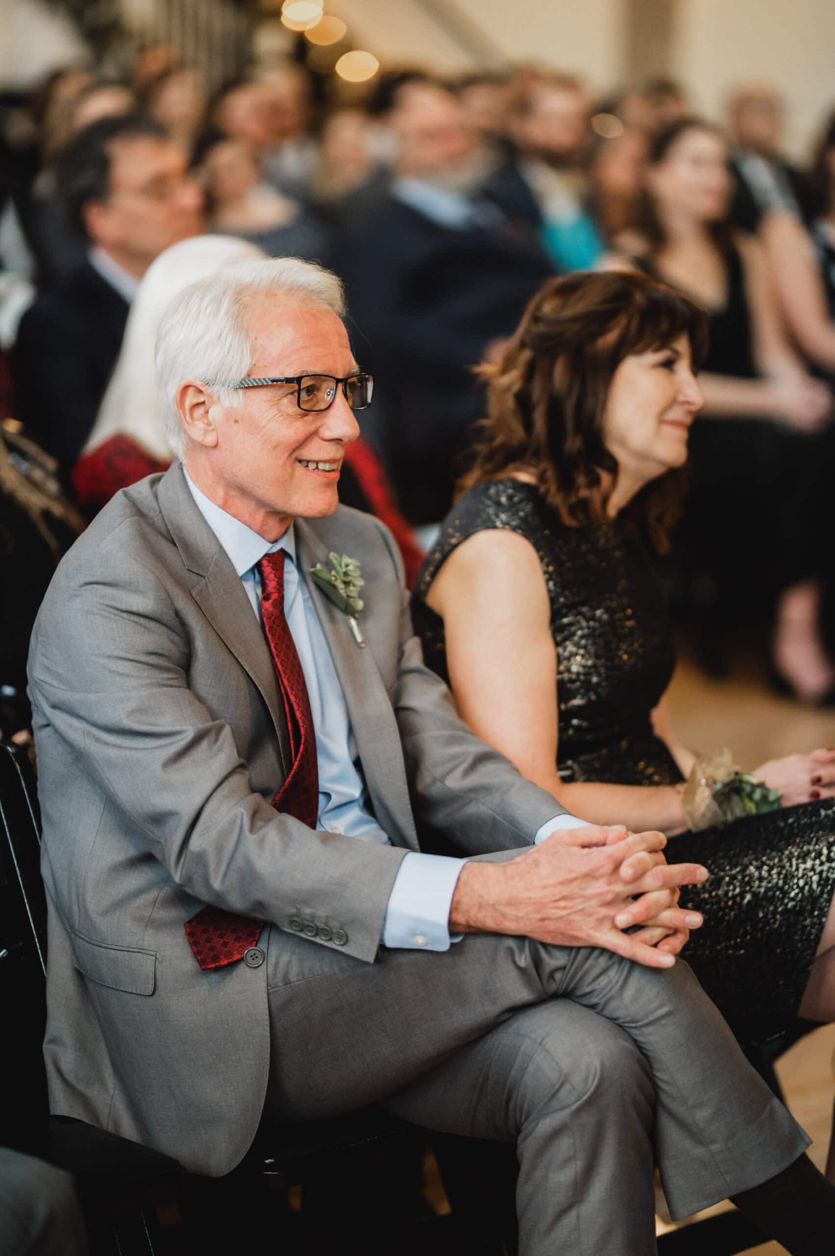 An elderly man in a gray suit and a woman in a black dress sit smiling at the crowded gathering, captured beautifully in wedding photos at The Evergreen.