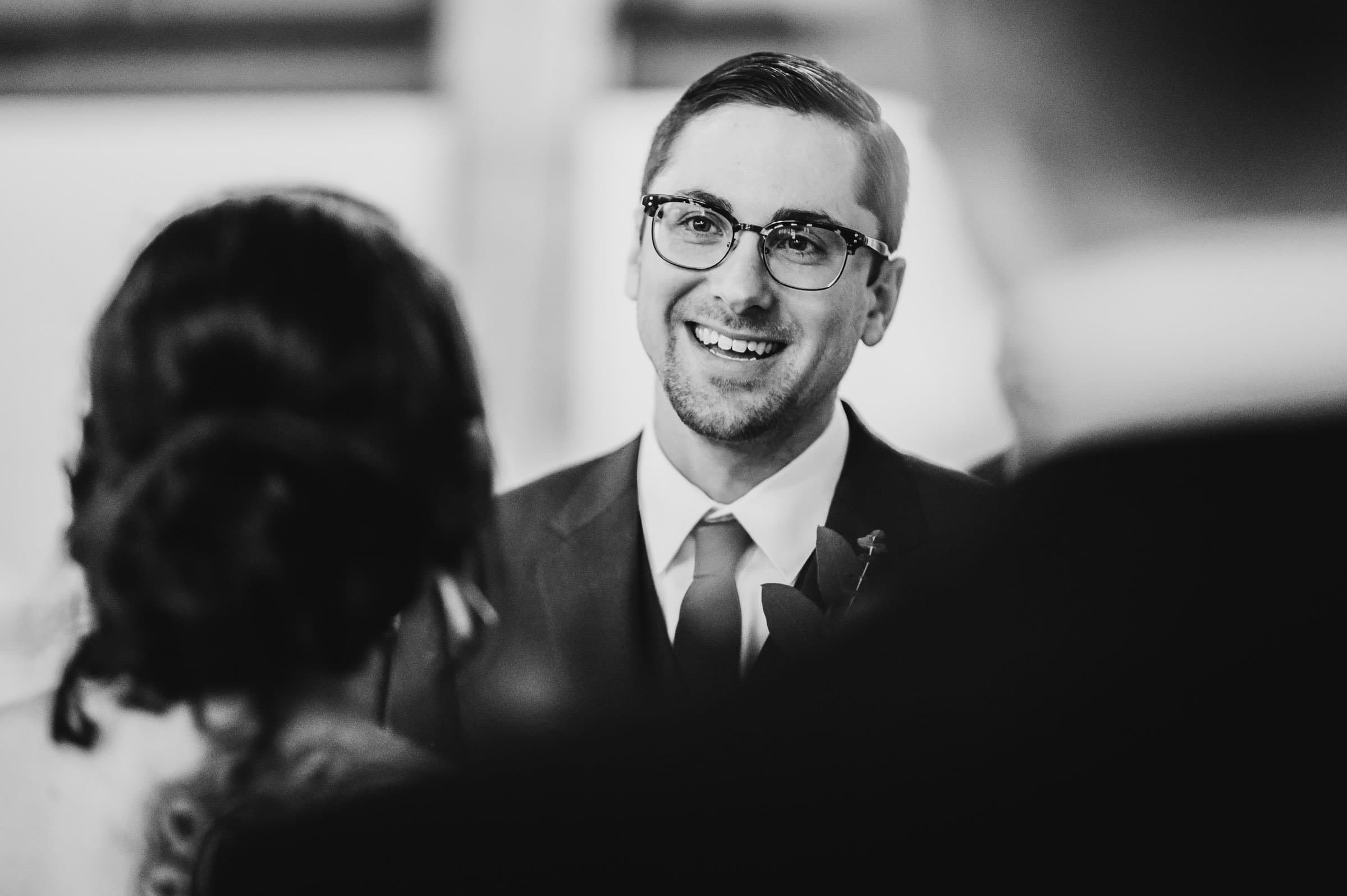 Man in glasses smiling at another person, dressed in a suit and tie, captured in a classic black-and-white setting. The photo exudes the charm of wedding photos at The Evergreen, epitomizing timeless elegance.