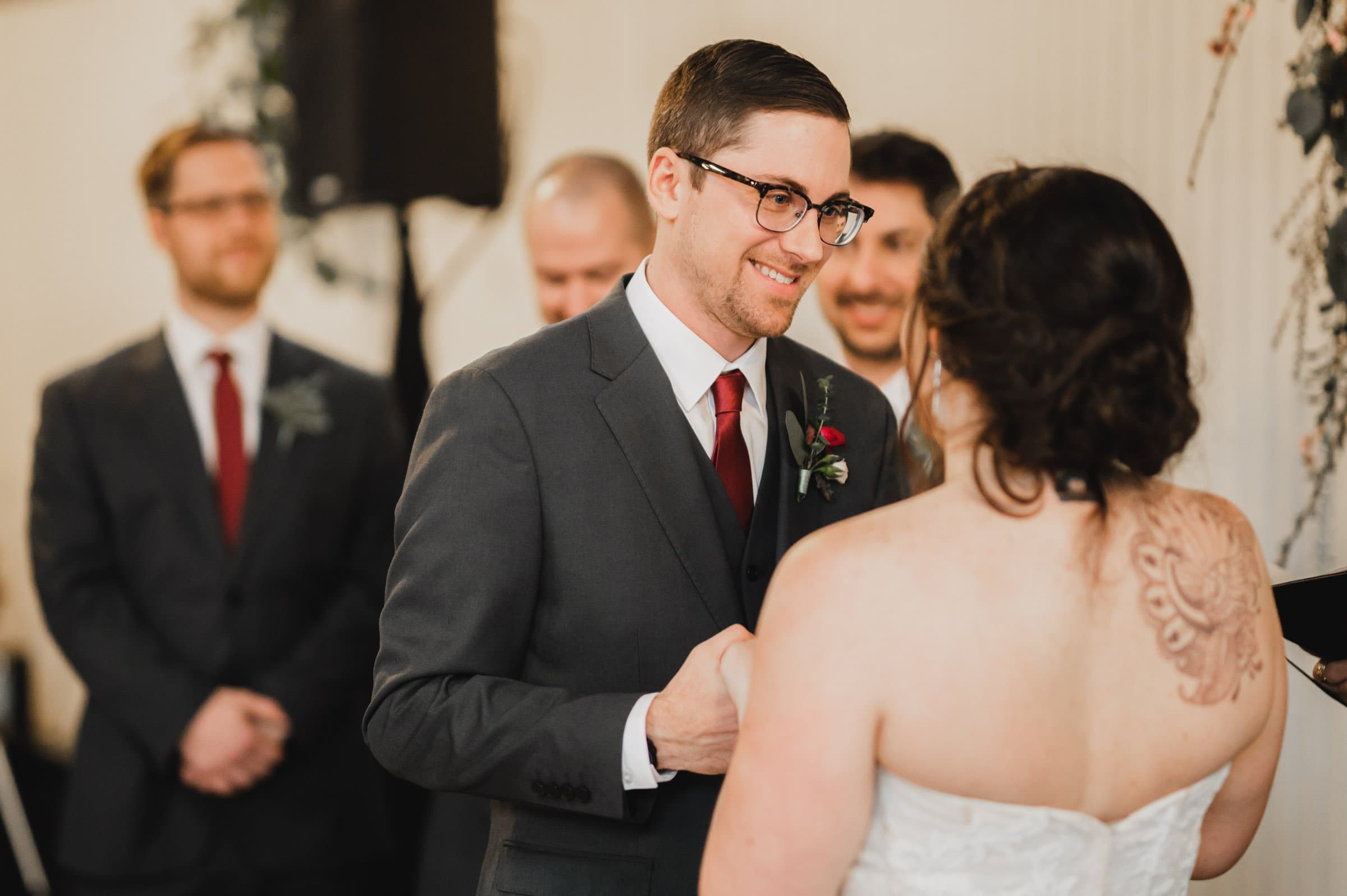 A groom smiles at his bride during their wedding ceremony, captured beautifully in the wedding photos at The Evergreen, with groomsmen standing in the background.
