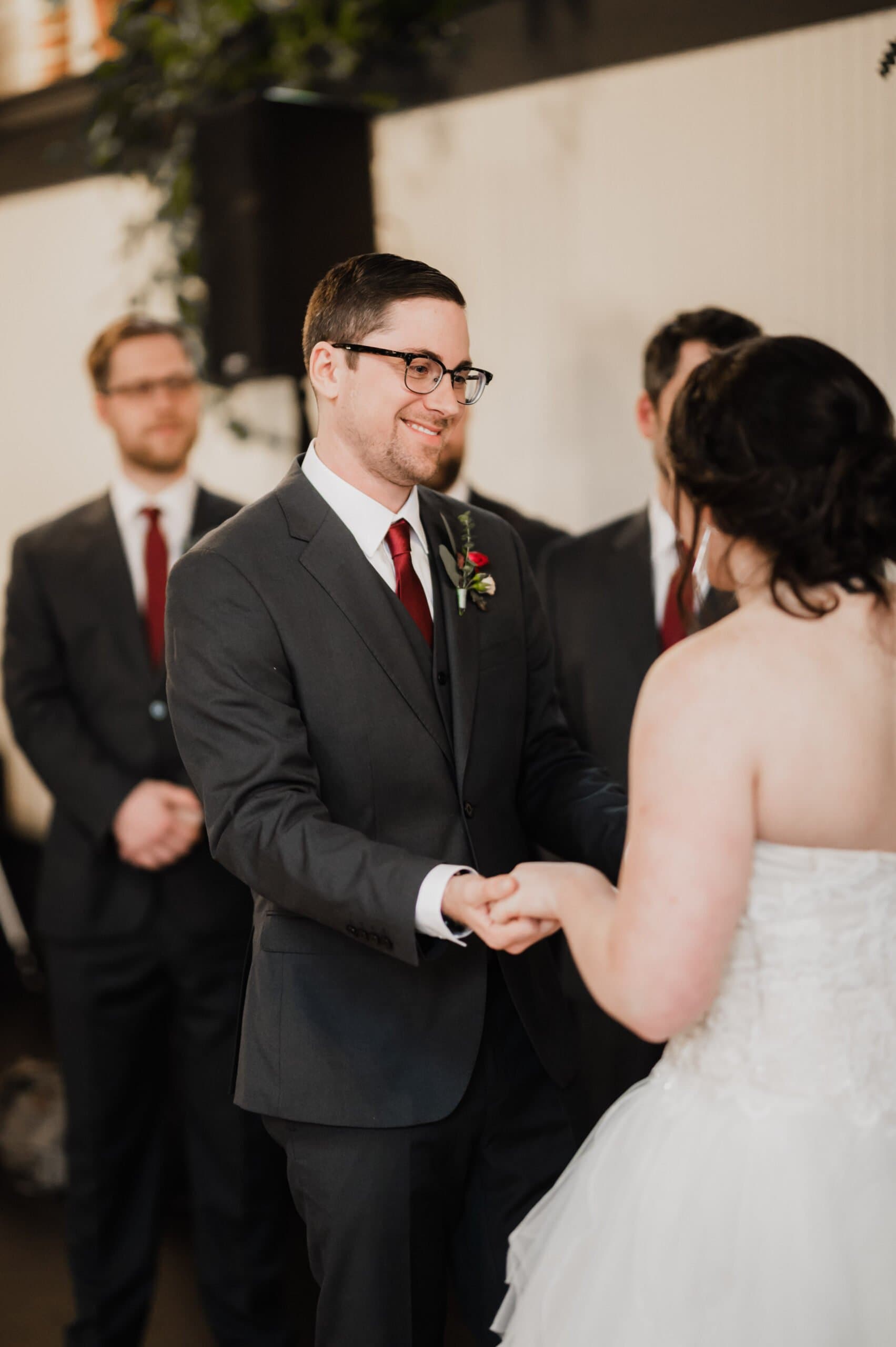 Groom in glasses holds brides hands during the wedding ceremony at The Evergreen, both smiling. Groomsmen in suits stand behind them.