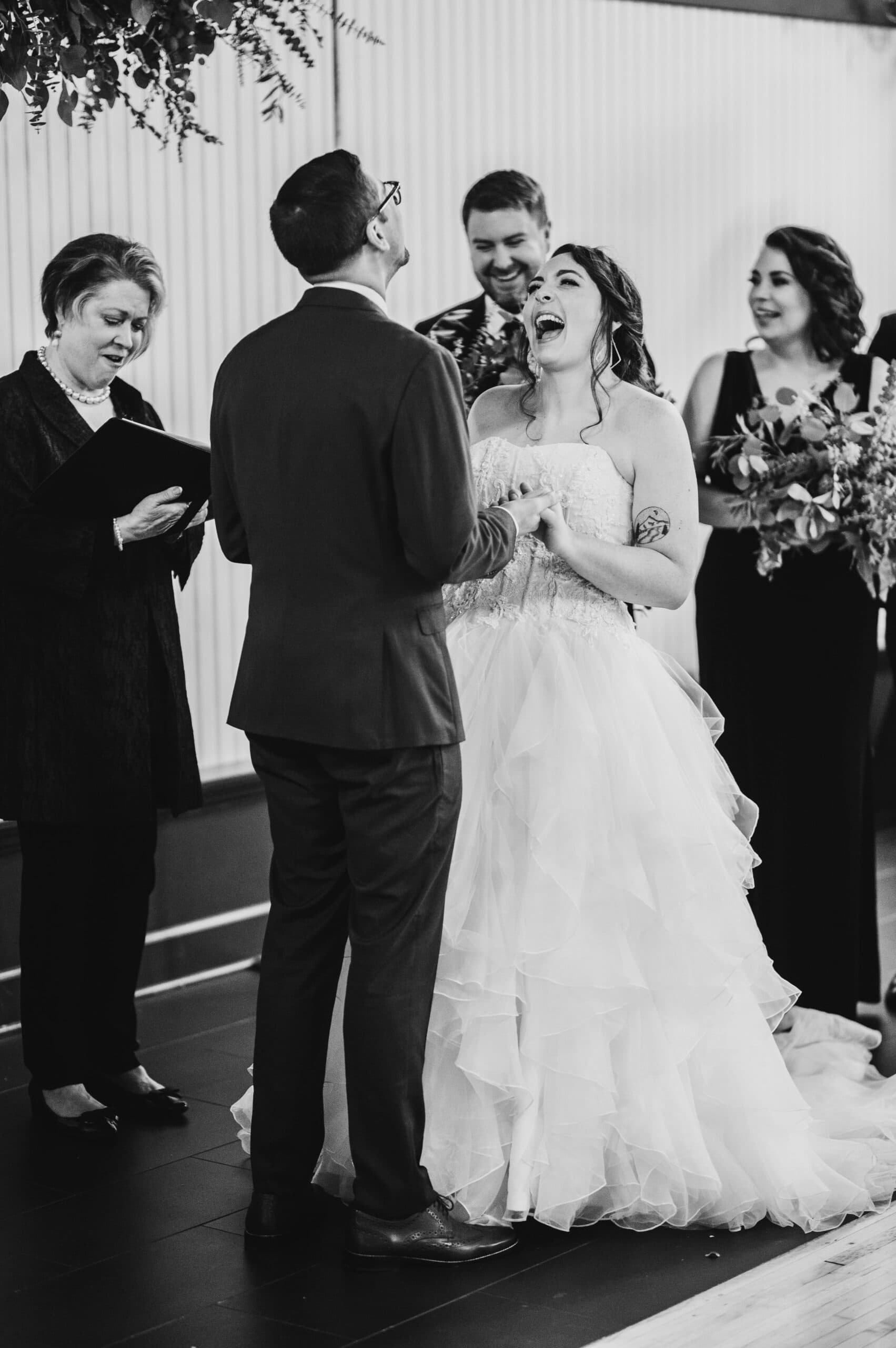 The bride and groom share a joyful laugh during their ceremony, with the officiant and bridesmaids adding to the delightful scene. This magical moment, captured as part of their wedding photos at The Evergreen, embodies love and happiness.