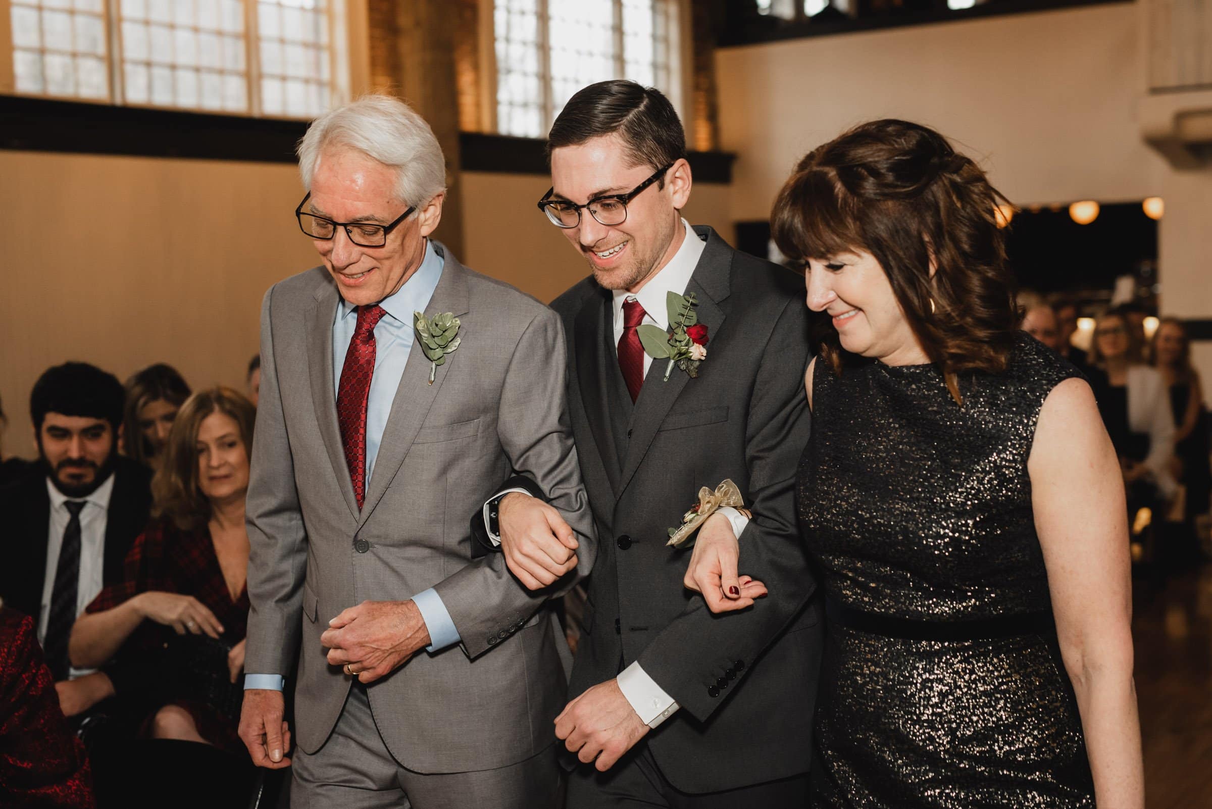 A groom walks down the aisle with an older man and a woman, all smiling warmly at the beautifully lit venue, capturing perfect wedding photos at The Evergreen.