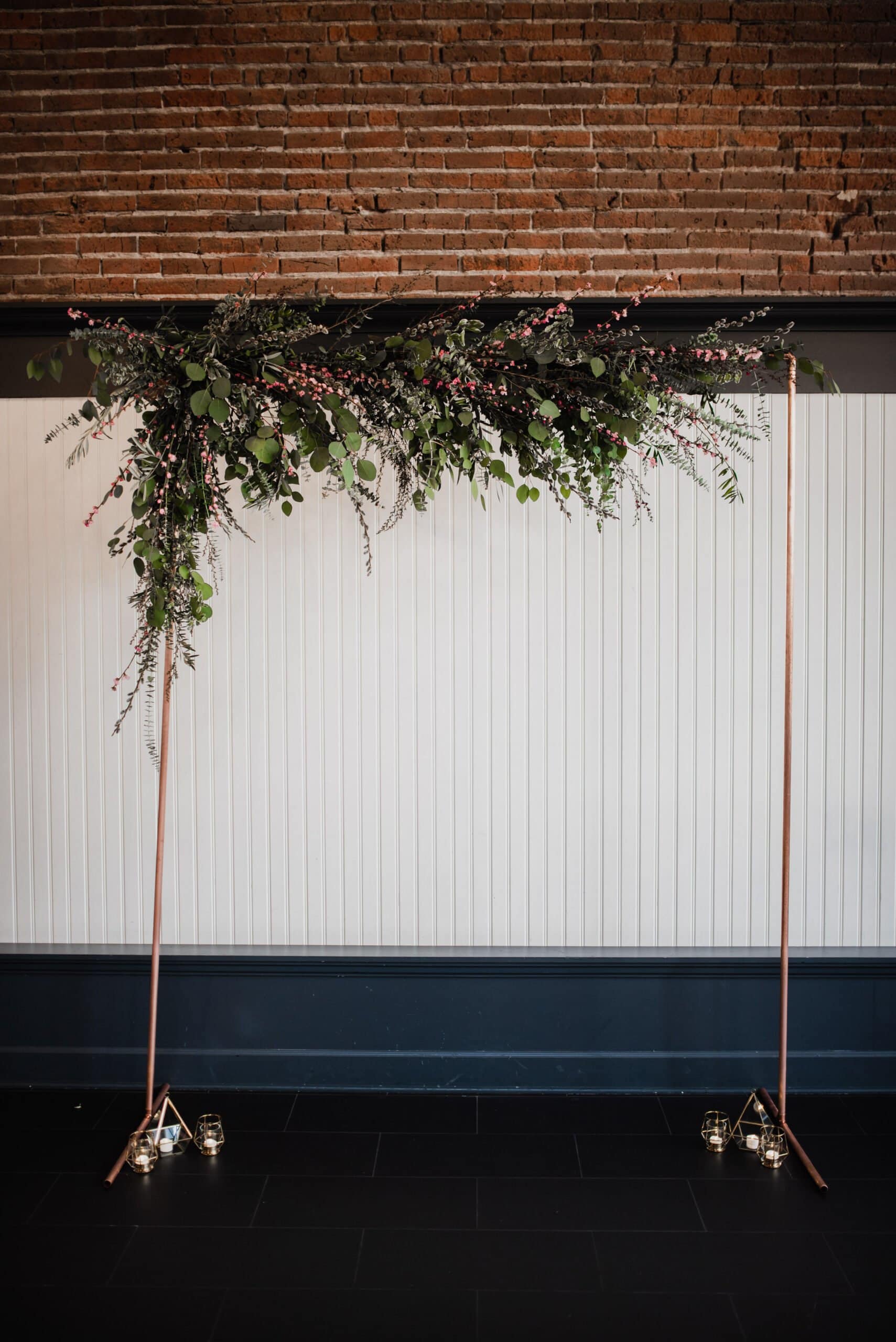 A stunning bronze wedding arch adorned with lush greenery and pink flowers stands elegantly against a brick and white paneled wall, perfect for capturing unforgettable wedding photos at The Evergreen.