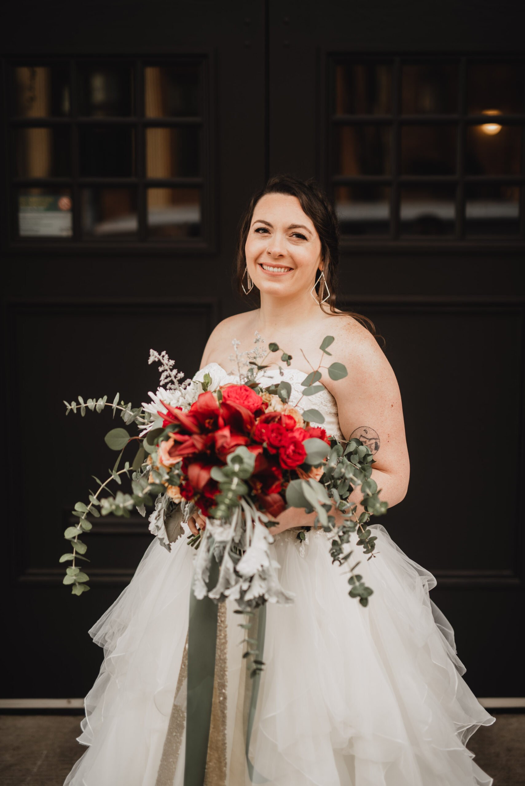 The bride in a white dress holds a bouquet of red flowers, smiling elegantly in front of dark double doors, capturing stunning wedding photos at The Evergreen.