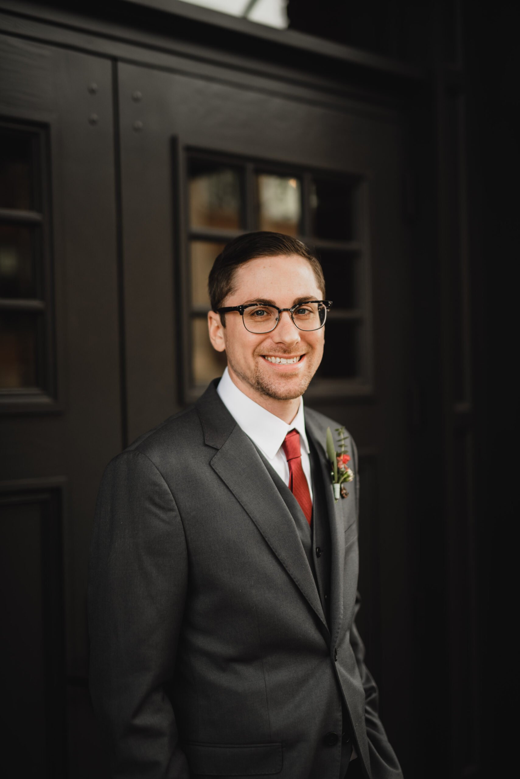 A man in a dark suit and red tie stands smiling in front of a dark door with window panels, captured beautifully like wedding photos at The Evergreen.