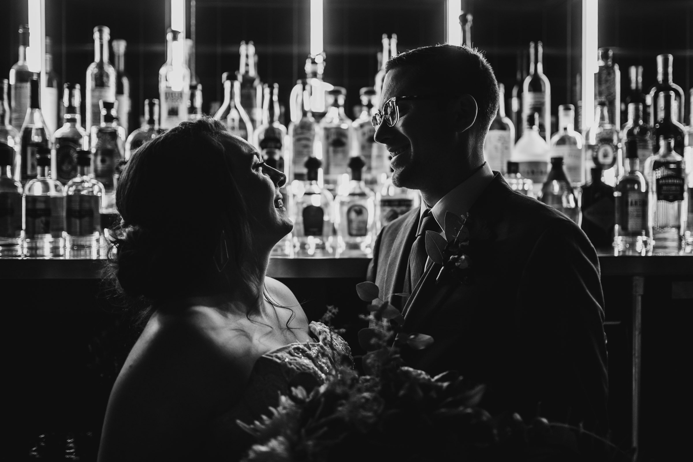 In a dimly lit bar adorned with illuminated bottles, the couple shares a tender smile during their wedding portraits at Voysey before they embark on their special day at The Evergreen.