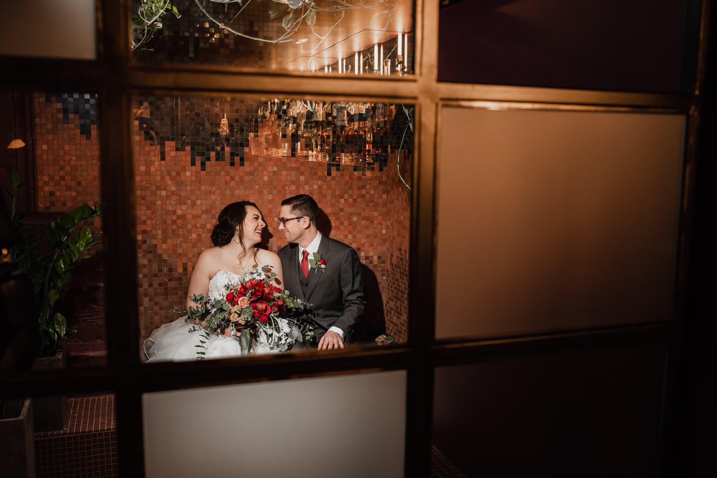 The bride and groom share a joyous smile in their wedding portraits at Voysey, surrounded by the cozy warmth of their venue, with a bouquet of red flowers hinting at their upcoming celebration at The Evergreen.