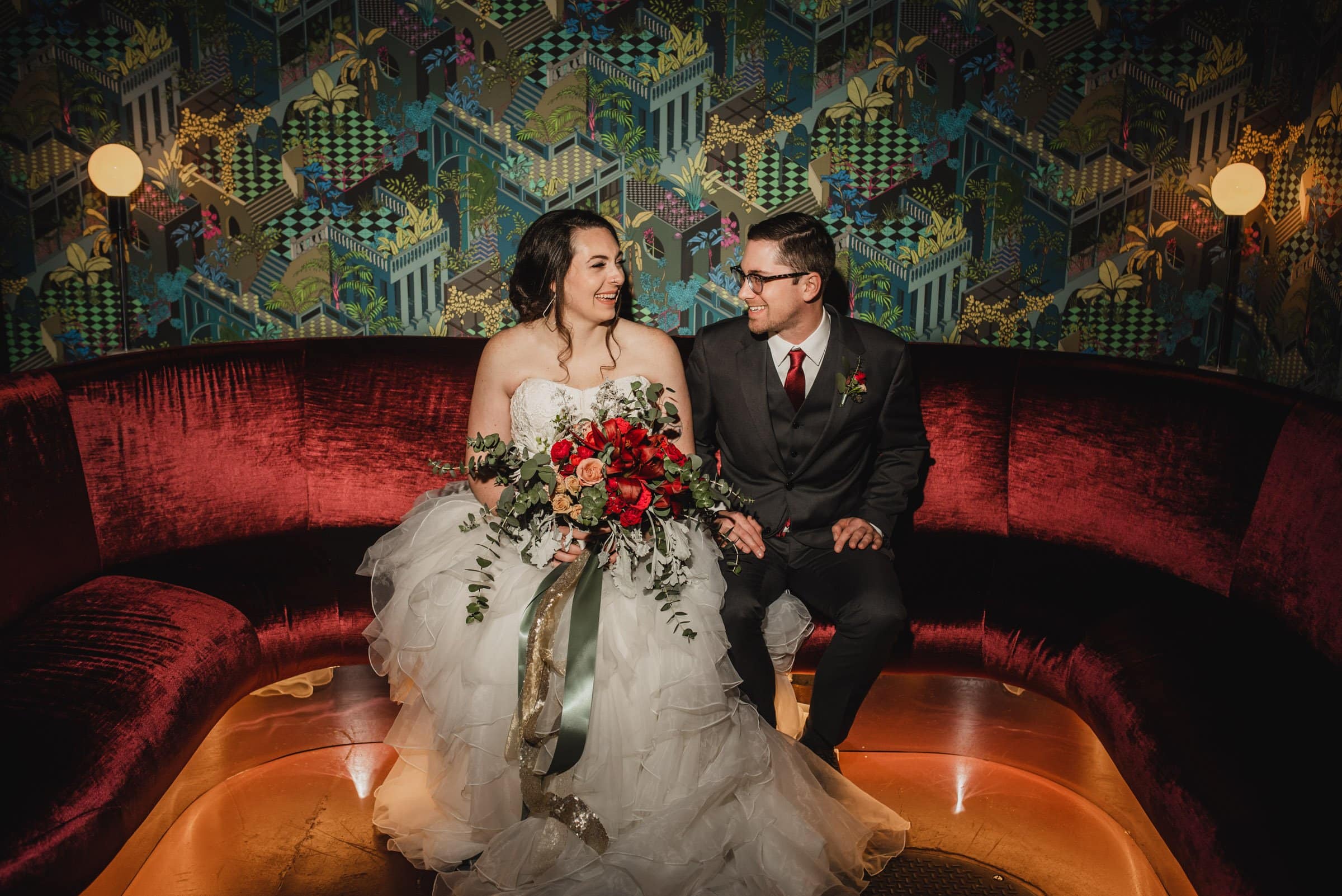 The bride and groom, moments before their wedding at The Evergreen, sit on a red velvet couch at Voysey, smiling at each other against a vibrant, patterned wall.