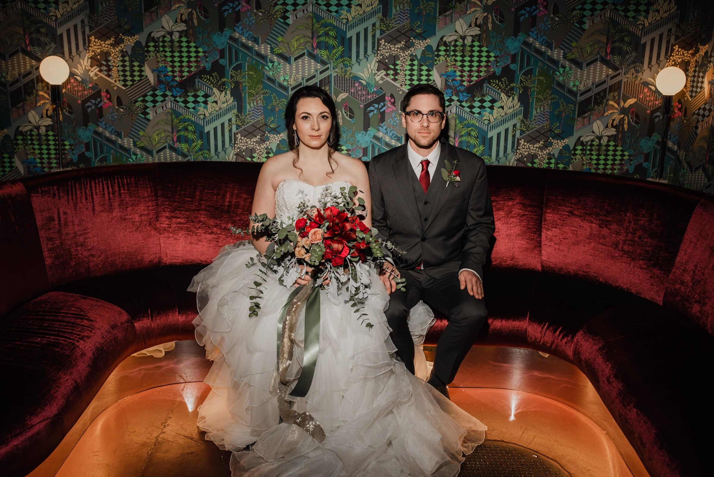 The bride and groom are elegantly seated on a red velvet couch adorned with floral wallpaper, holding a vibrant red bouquet. These charming wedding portraits at Voysey capture their joy before their wedding at The Evergreen.