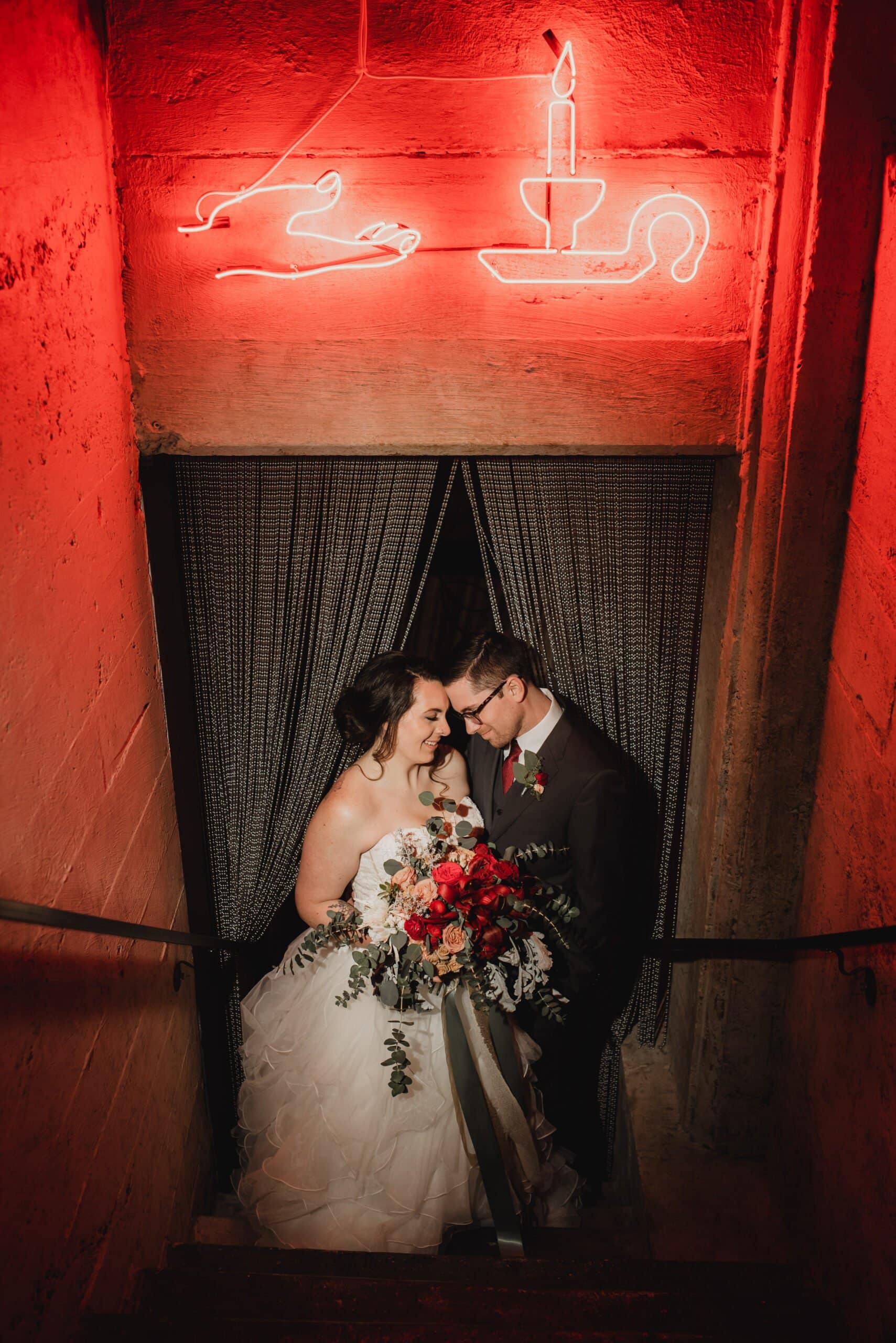 The bride and groom smile and embrace on the dimly lit stairs under a neon candle sign, capturing stunning wedding portraits at Voysey before their celebration at The Evergreen.