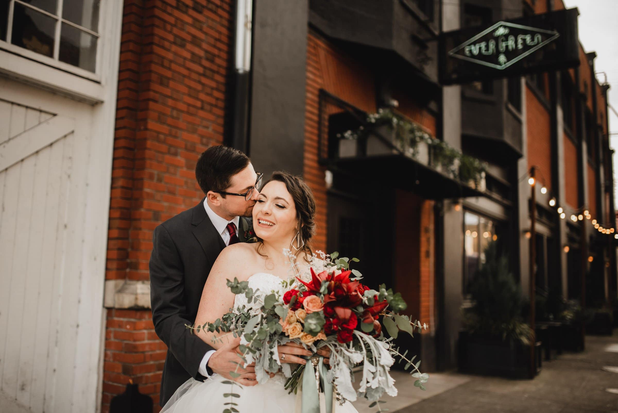 The bride and groom embrace joyfully outside a brick building, holding a bouquet of red and green flowers, capturing the magic of their special day with wedding photos at The Evergreen.
