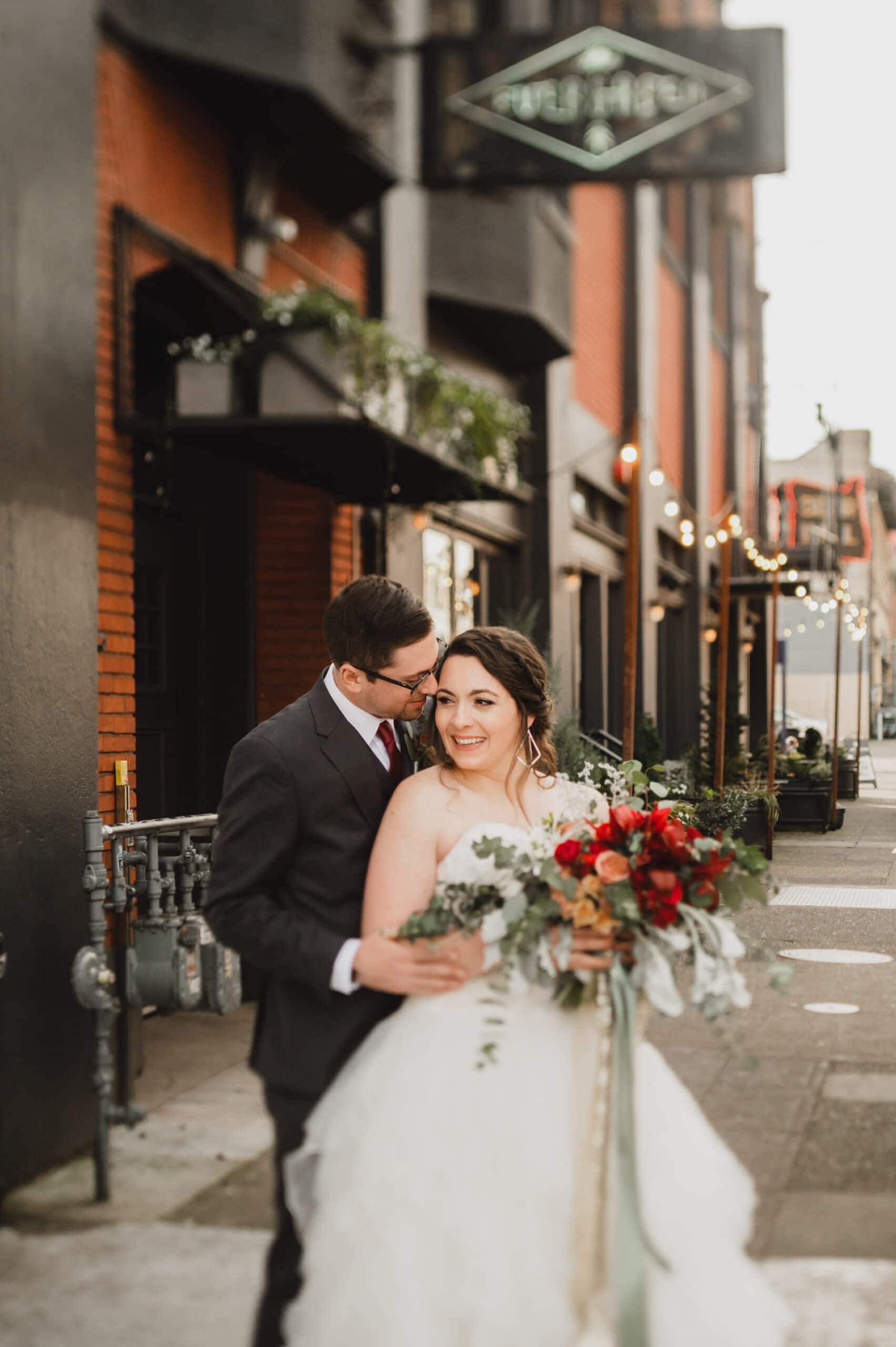 Bride and groom smiling on a city street, holding a bouquet with red and white flowers, capturing their special day with wedding photos at The Evergreen.