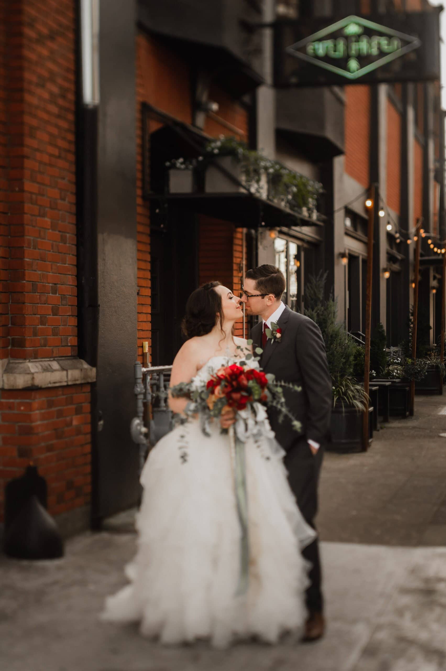 The bride and groom share a kiss on a city sidewalk, holding a bouquet in front of a brick building, capturing beautiful wedding photos at The Evergreen.