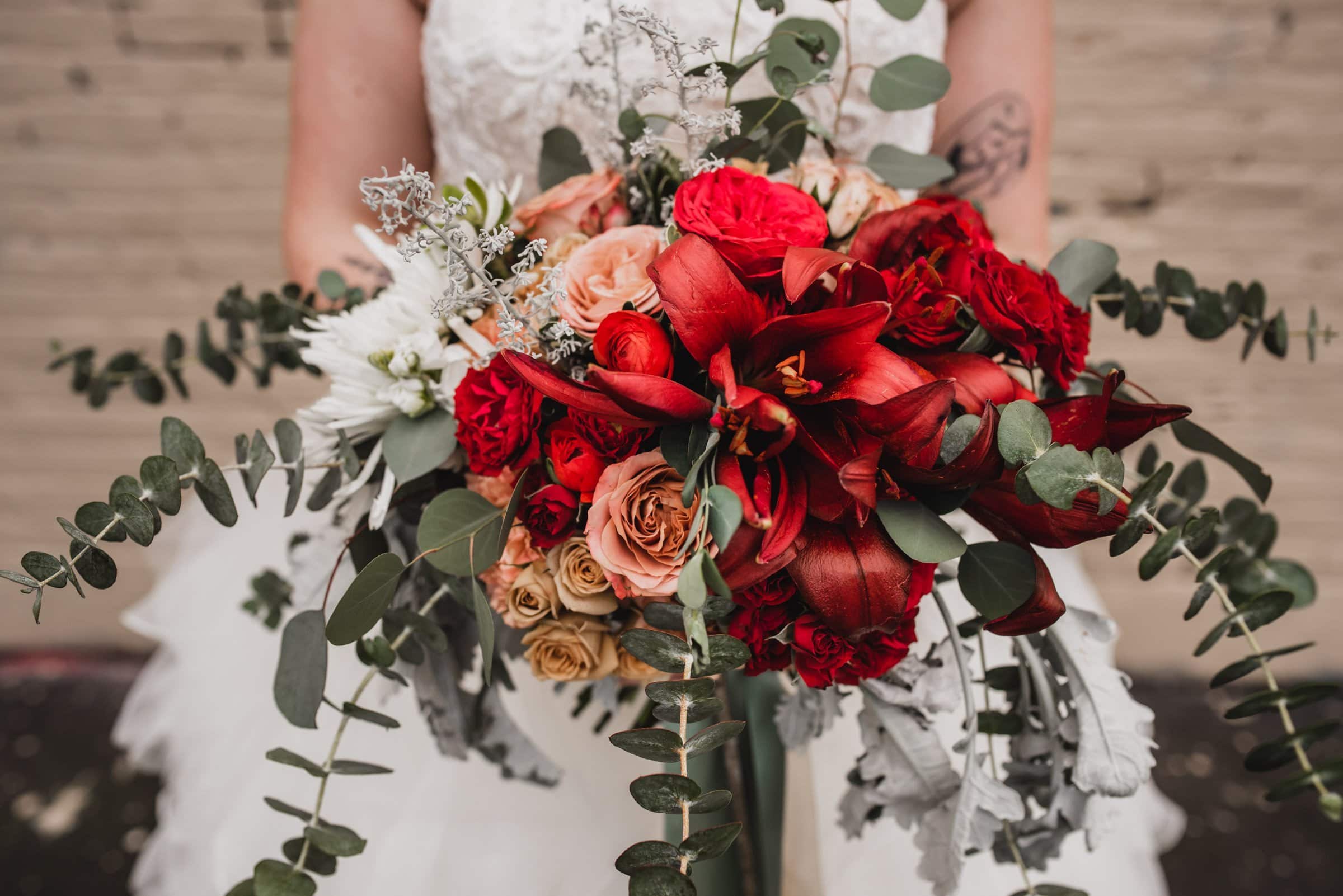 The bride, holding a bouquet of red and peach flowers complemented by lush greenery and eucalyptus, poses elegantly for wedding photos at The Evergreen.