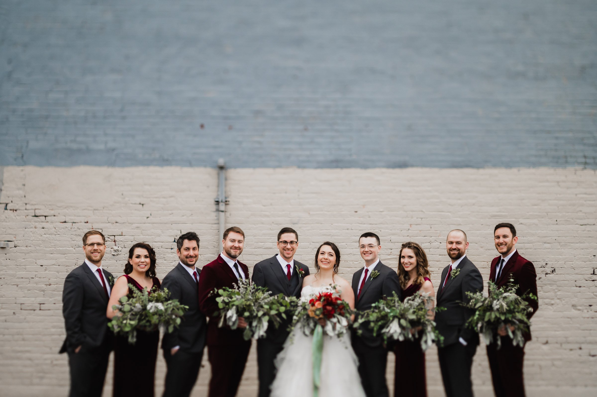 The bridal party is beautifully arranged against a brick wall, with the bride in white and bridesmaids in dark dresses, while the groomsmen look sharp in suits. This charming scene captures the elegance of wedding photos at The Evergreen.