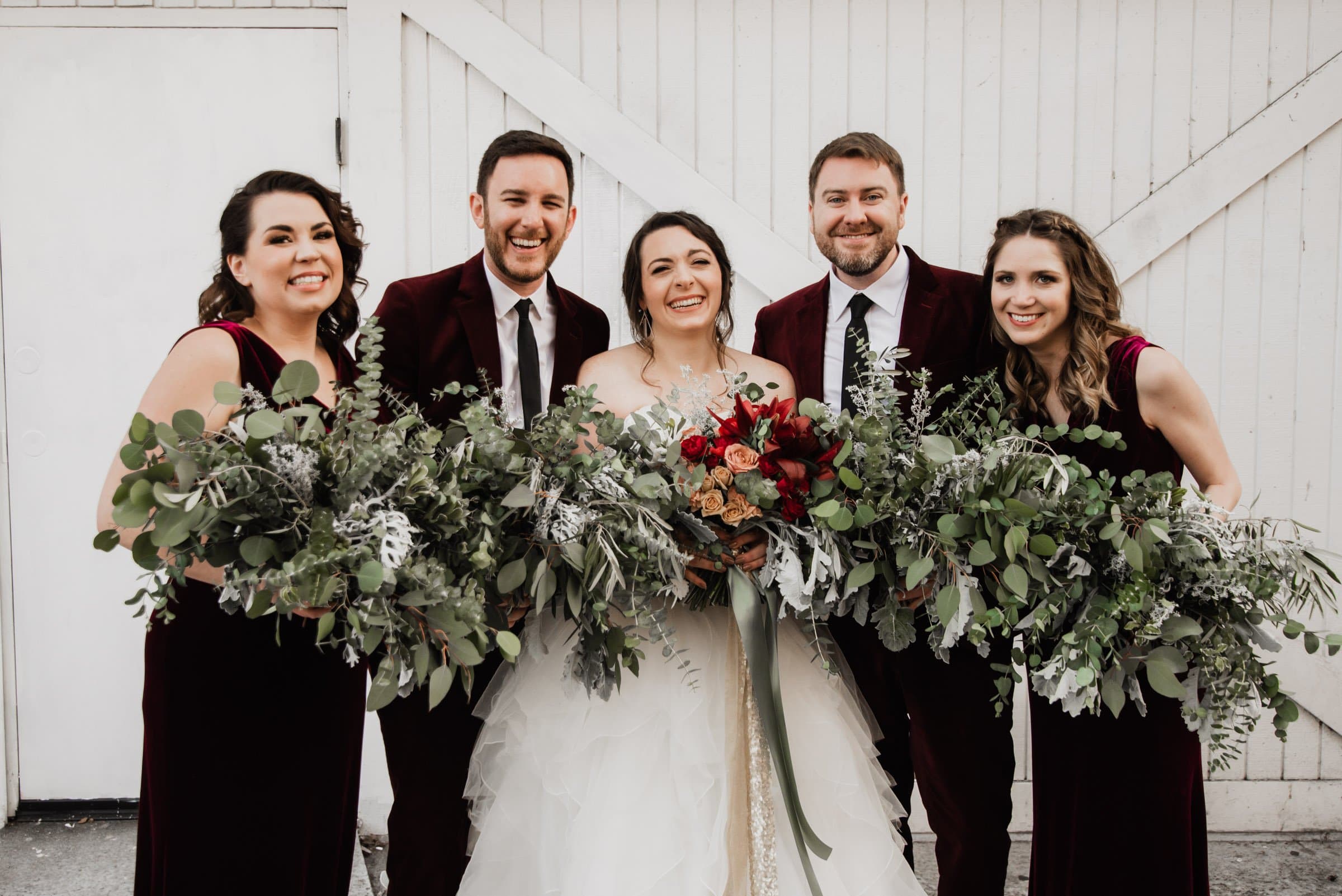The bride and wedding party are all smiles as they hold vibrant bouquets in front of a white wooden wall, capturing perfect wedding photos at The Evergreen.