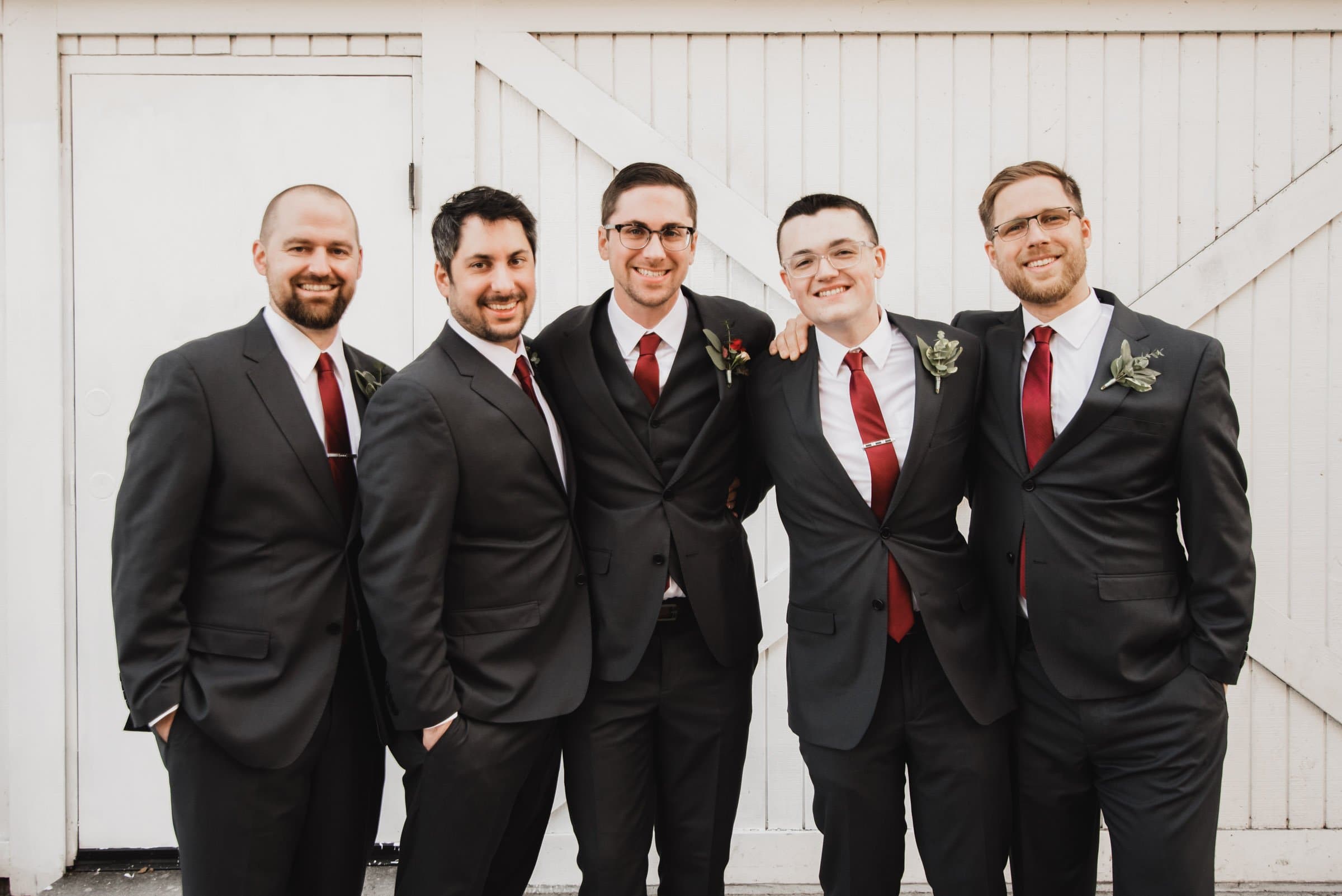 Five men in suits with red ties stand smiling in front of a white wooden wall, their camaraderie captured beautifully. Its like a candid moment from wedding photos at The Evergreen, where joy and style meet effortlessly.