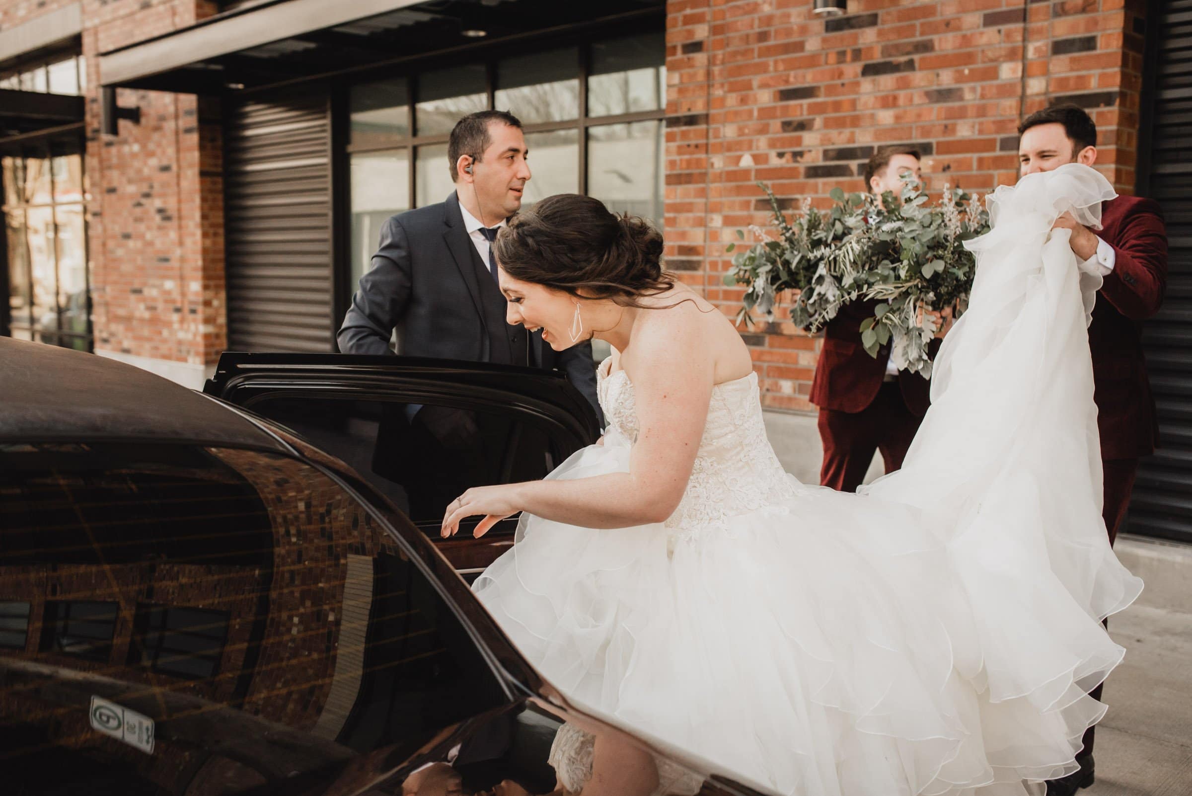 Bride entering car with help; two men assist while another holds her bouquet. A first look at a nearby apartment building provides a charming backdrop before the wedding at The Evergreen.