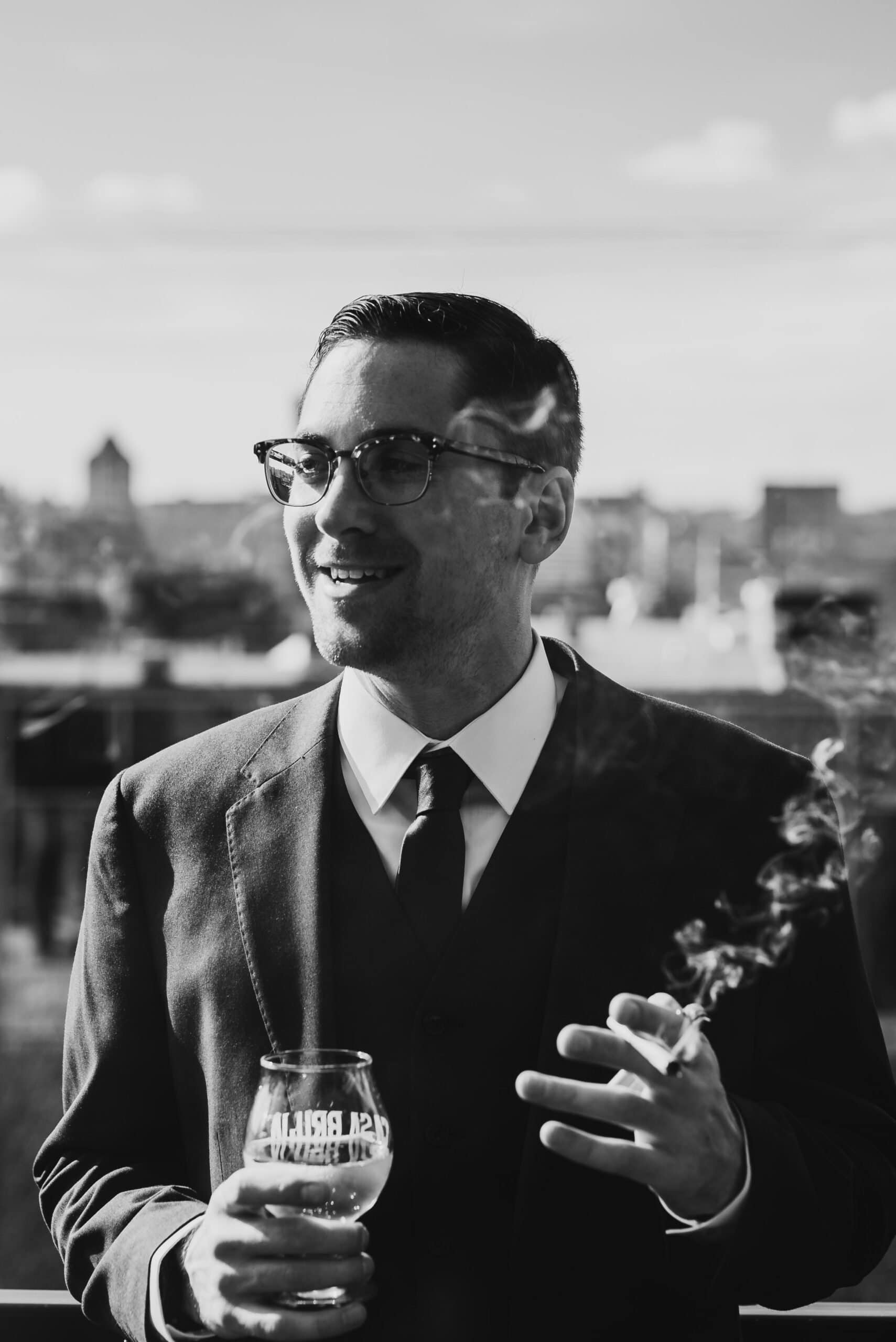 Man in a suit with glasses, getting ready at a nearby apartment for a wedding at The Evergreen, holds a drink and smokes as he stands outside with the cityscape in the background. Black and white photo.