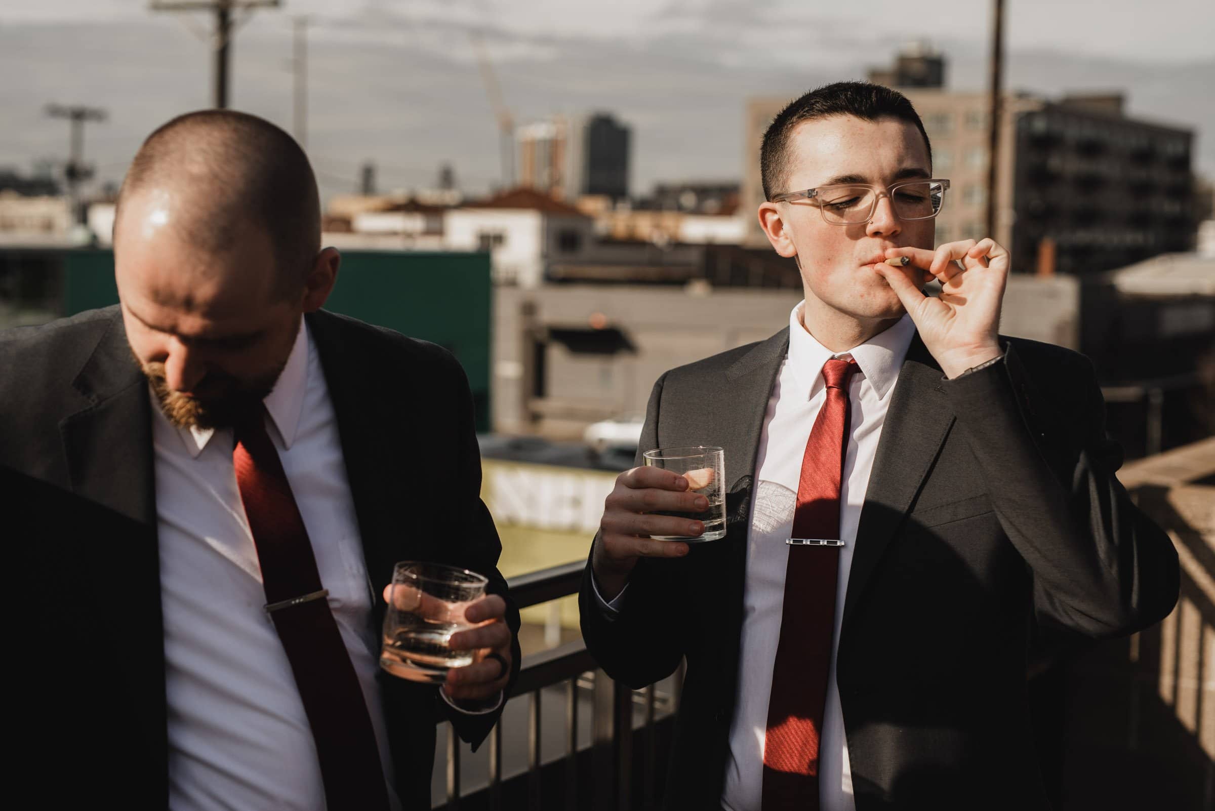 Two men in suits with red ties, one smoking a cigar and the other holding a drink, stand on a rooftop, having just gotten ready at a nearby apartment for a wedding at The Evergreen.