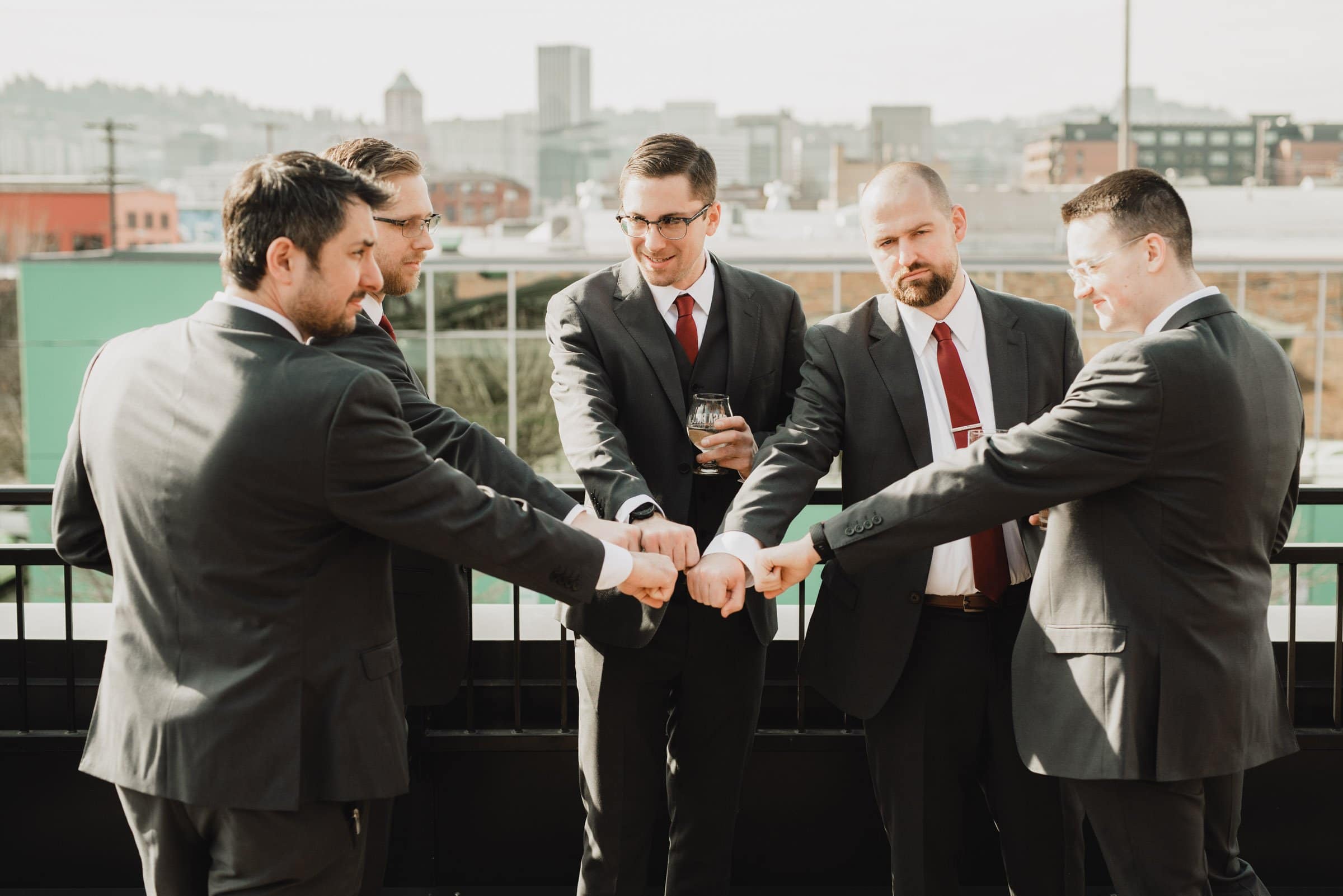 Five men in suits, fresh from getting ready for a wedding at a nearby apartment, do a group fist bump on a rooftop with the cityscape and The Evergreen in the background.