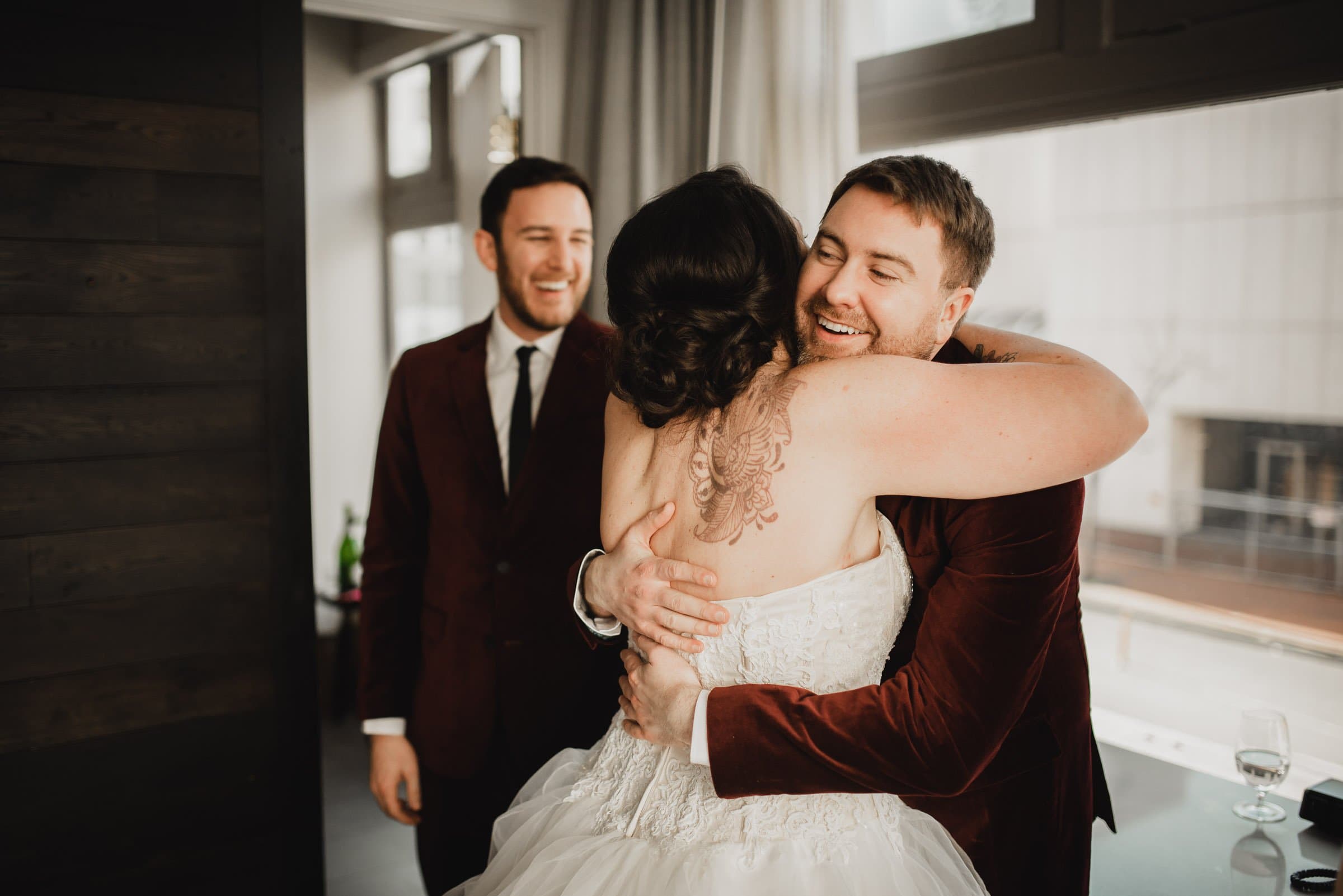 Bride in white dress hugs groom in maroon jacket while a smiling friend looks on in the bright room of the Hi-Lo Hotel.