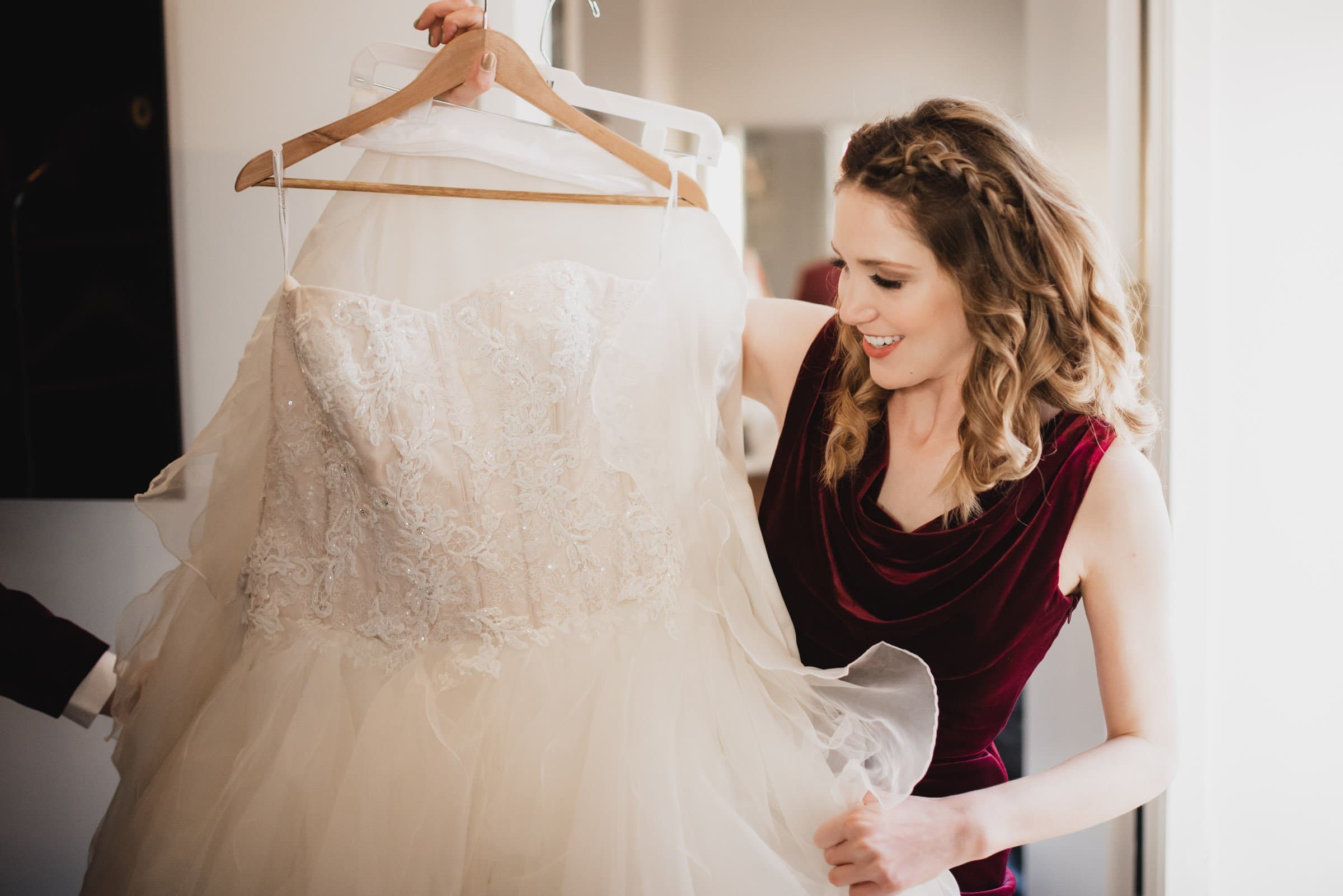 A woman in a burgundy dress stands indoors at the Hi-Lo Hotel, smiling as she holds a wedding gown on a hanger.