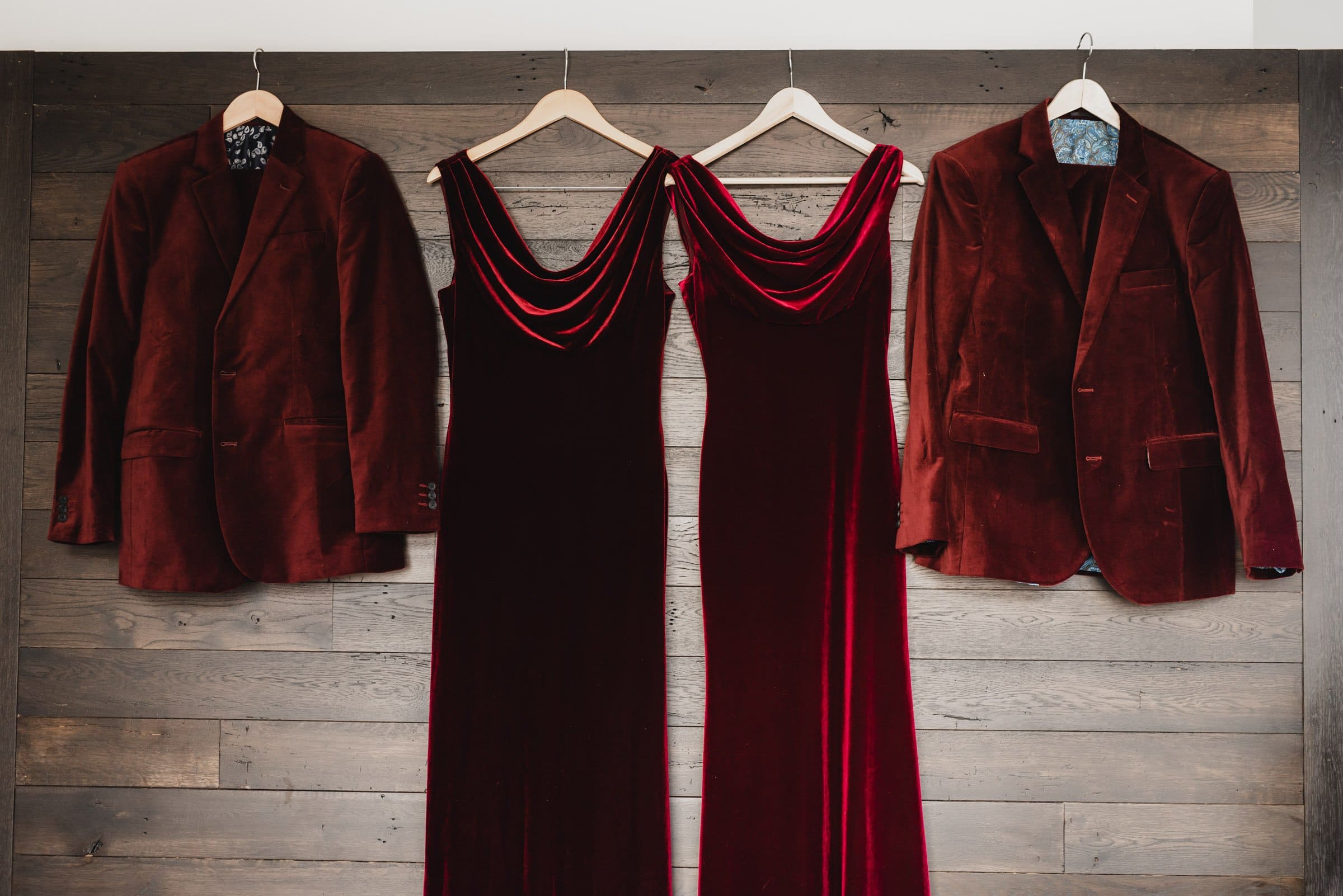 Two red velvet dresses and two jackets hang on wooden hangers against a rustic wooden wall, perfect for getting ready at the Hi-Lo Hotel.