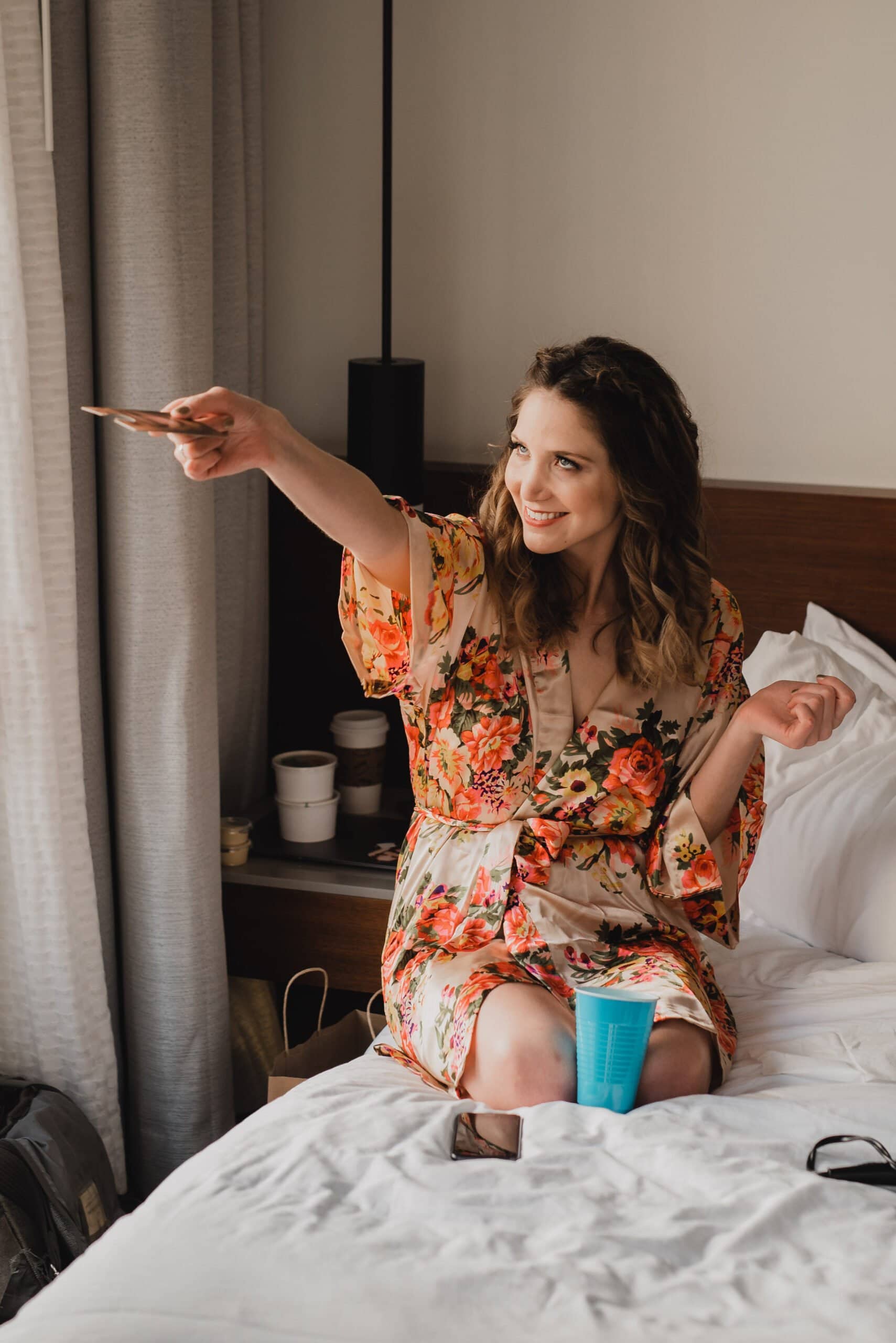 Woman in floral robe sits on bed at the Hi-Lo Hotel, smiling and pointing. A blue cup and sunglasses are beside her, capturing a moment of serene preparation.