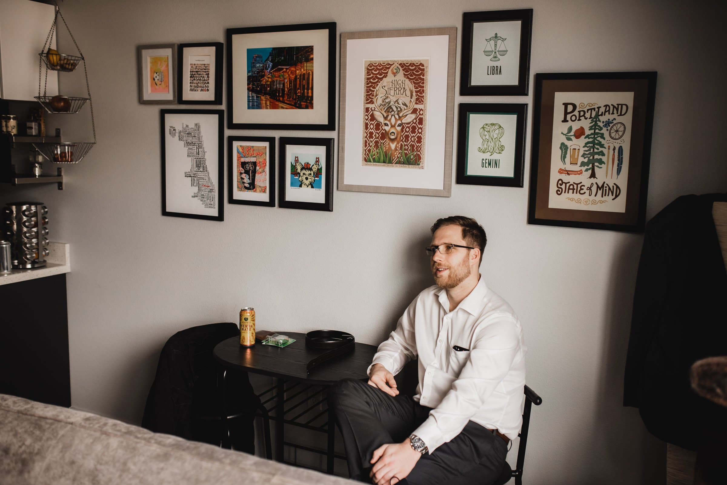 A man sits by a table with a drink, surrounded by framed artwork on the wall in a cozy room, contemplating his next move as he gets ready at a nearby apartment for a wedding at The Evergreen.