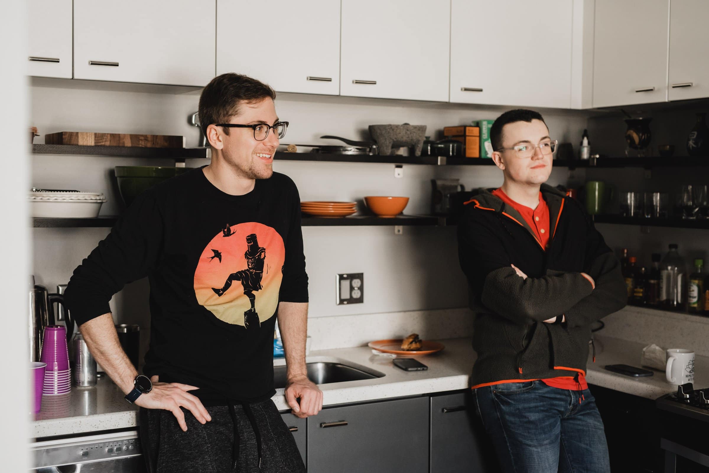 Two men in a kitchen, one leaning on the counter, both casually dressed and wearing glasses, are getting ready at a nearby apartment for a wedding at The Evergreen.