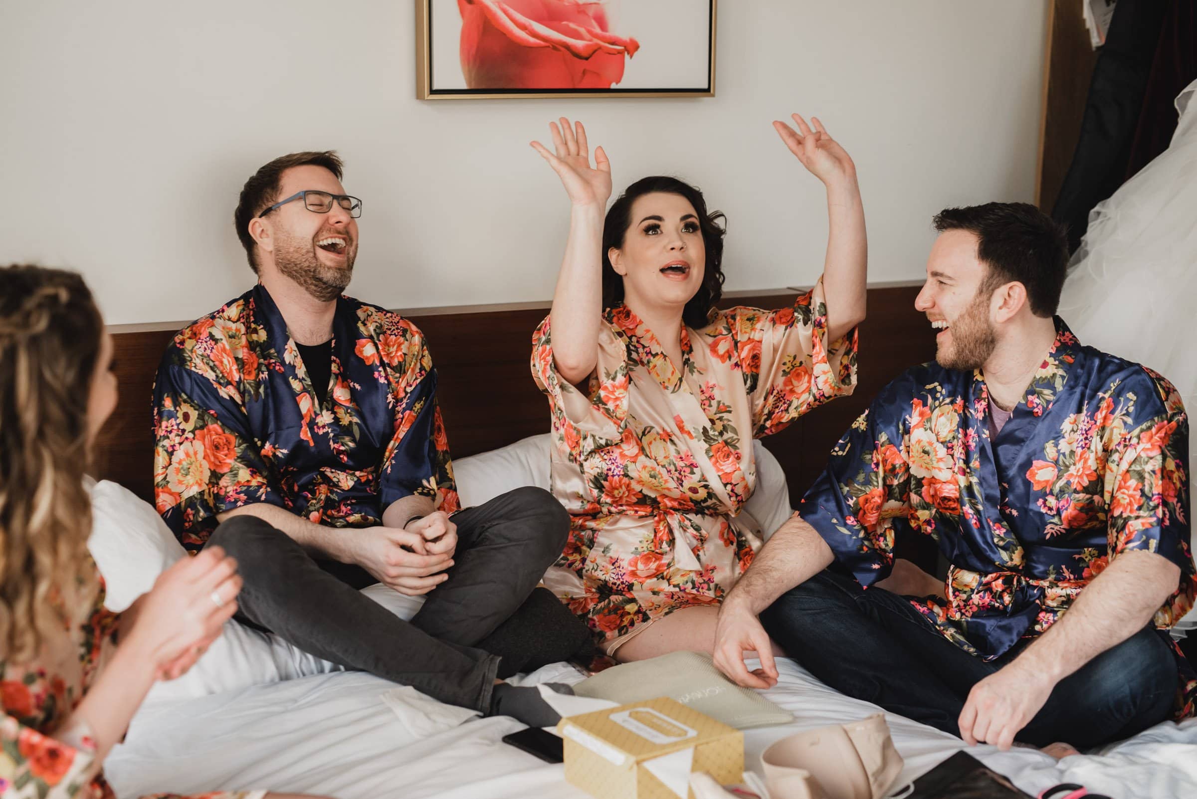 Four people in floral robes laugh and gesture happily while sitting on a bed, getting ready at the Hi-Lo Hotel.