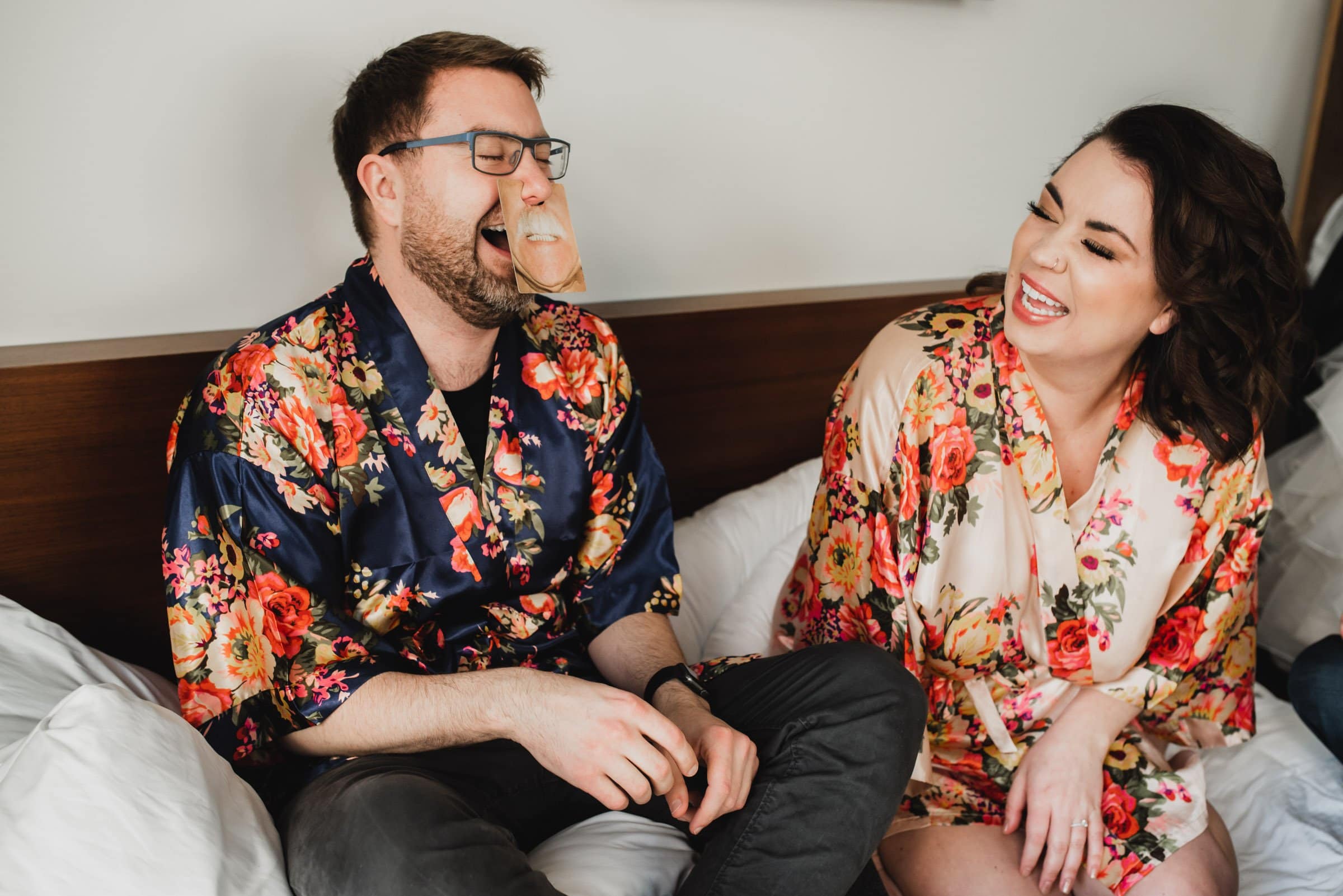 A man and woman in floral robes laugh together on a bed at the Hi-Lo Hotel; hes wearing glasses with a card attached.