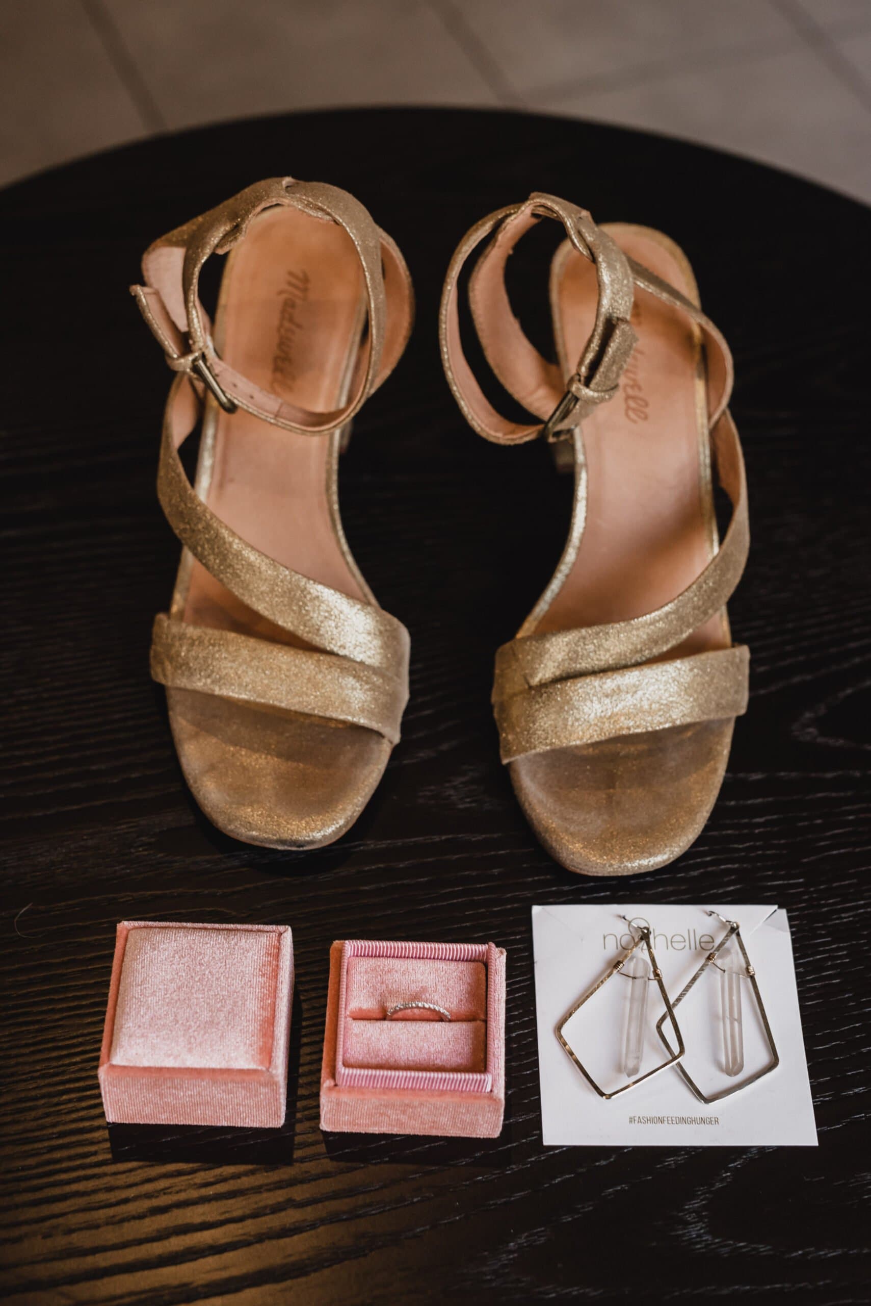 Gold high-heeled sandals paired with jewelry, including rings and geometric earrings, are arranged on a dark surface, perfect for getting ready at the Hi-Lo Hotel.