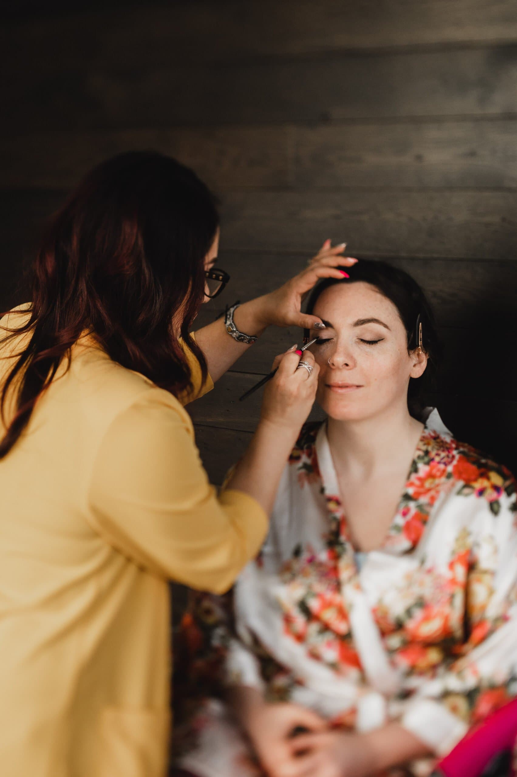 In a dark wood setting, a woman applies makeup to another wearing a floral robe, as they get ready at the Hi-Lo Hotel.