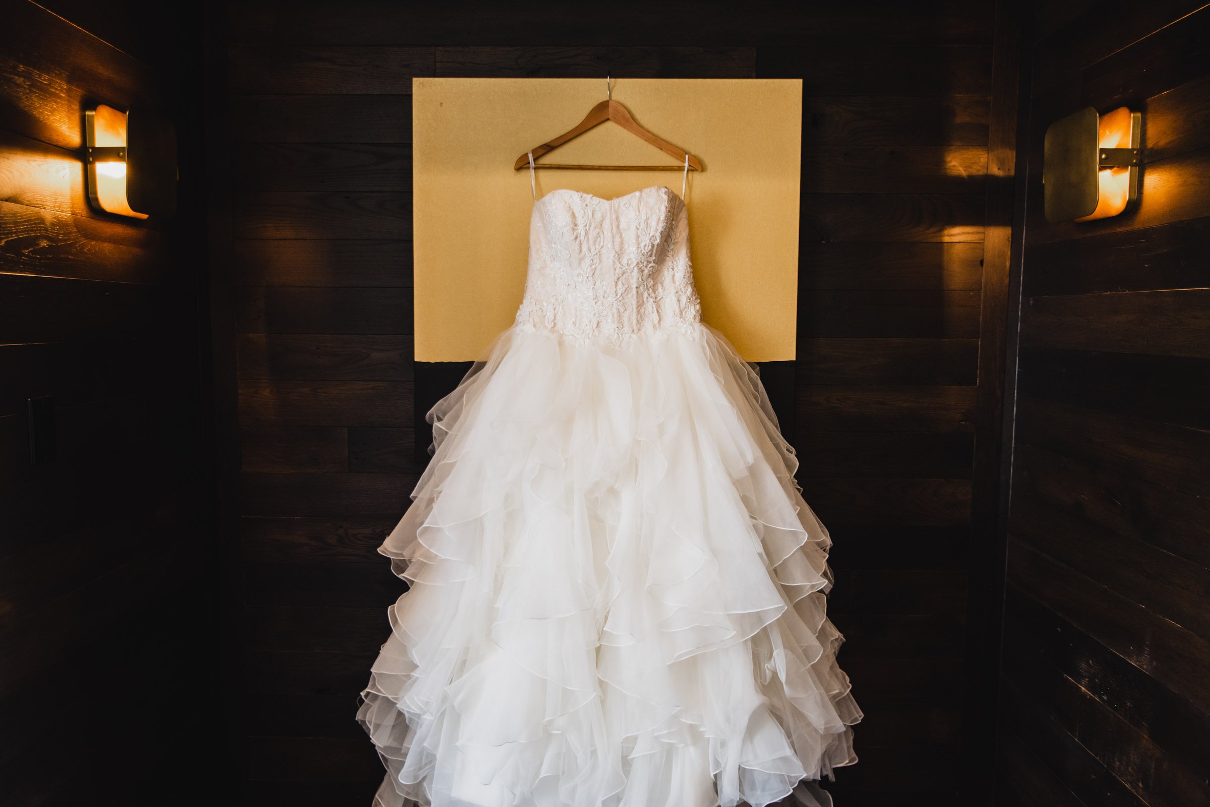 A white, strapless wedding dress hangs on a wooden hanger against a dark wooden wall at the Hi-Lo Hotel, illuminated by delicate wall lights.