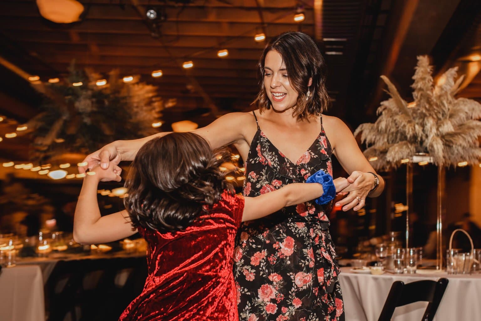 A woman in a floral dress dances with a child in a red dress under the warmly lit ambiance of a decorated room, capturing joyful moments that could belong to cherished wedding photos at Castaway Portland.