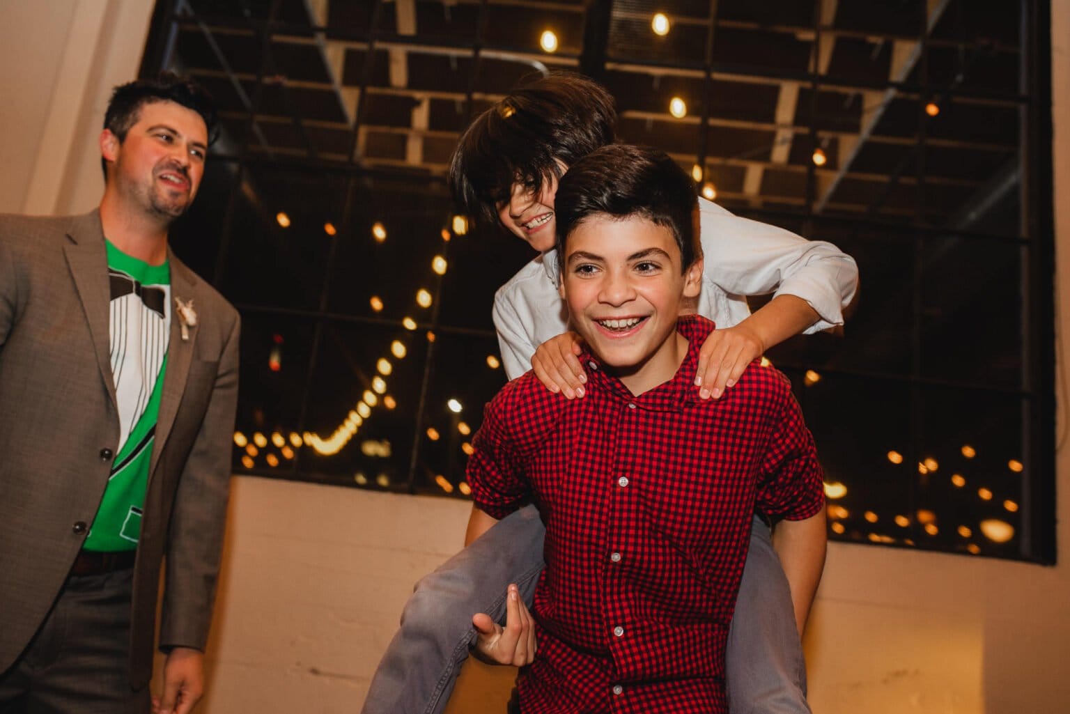In a festive indoor setting with lights glowing overhead, a person in a red shirt joyfully gives a piggyback ride to a smiling child. Nearby, another person stands, capturing the spirit found in wedding photos at Castaway Portland.