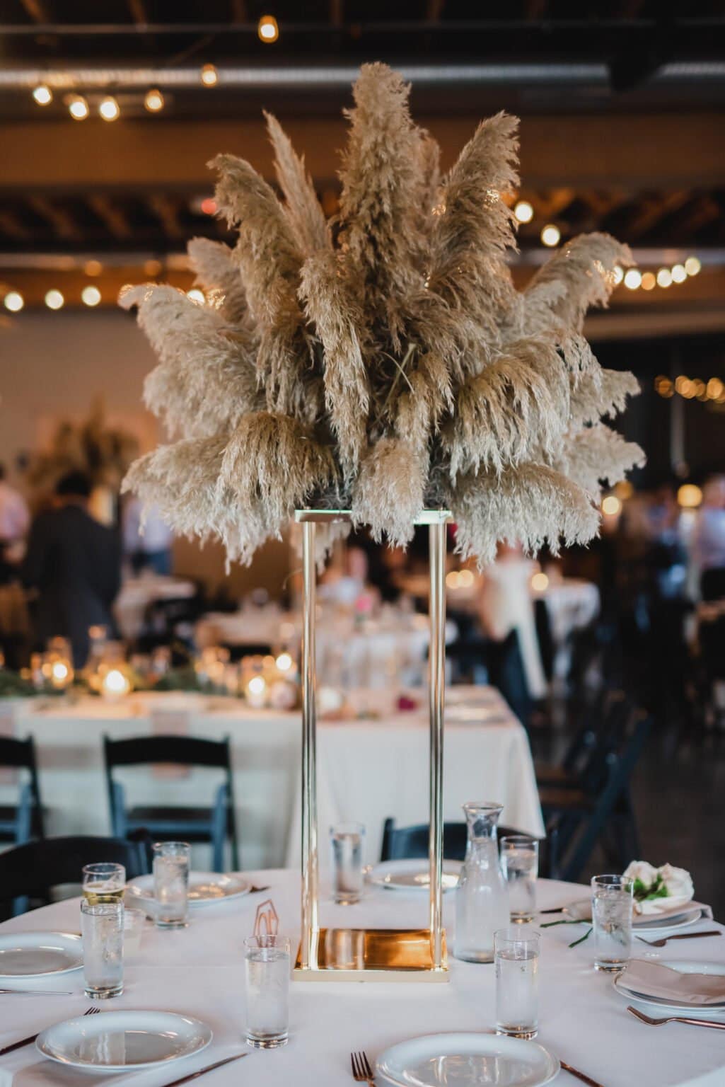 A tall centerpiece with pampas grass graces the dining table, surrounded by glasses and plates, creating a perfect setting for wedding photos at Castaway Portland. Warm lighting in the background adds to the inviting ambiance.