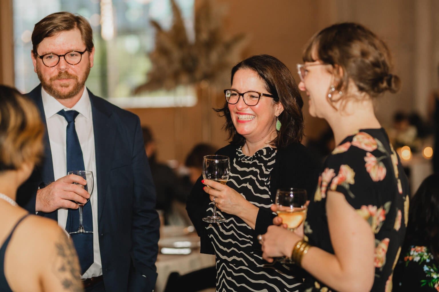 Three people are socializing at an event, holding wine glasses and smiling, dressed in formal attire. This moment captures the elegance of wedding photos at Castaway Portland.