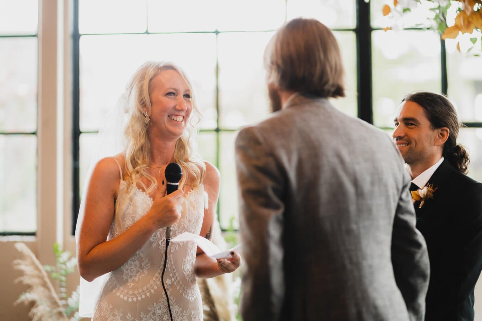 During the wedding ceremony at Castaway Portland, the bride smiled warmly with a microphone in hand as two guests faced her in the bright room.