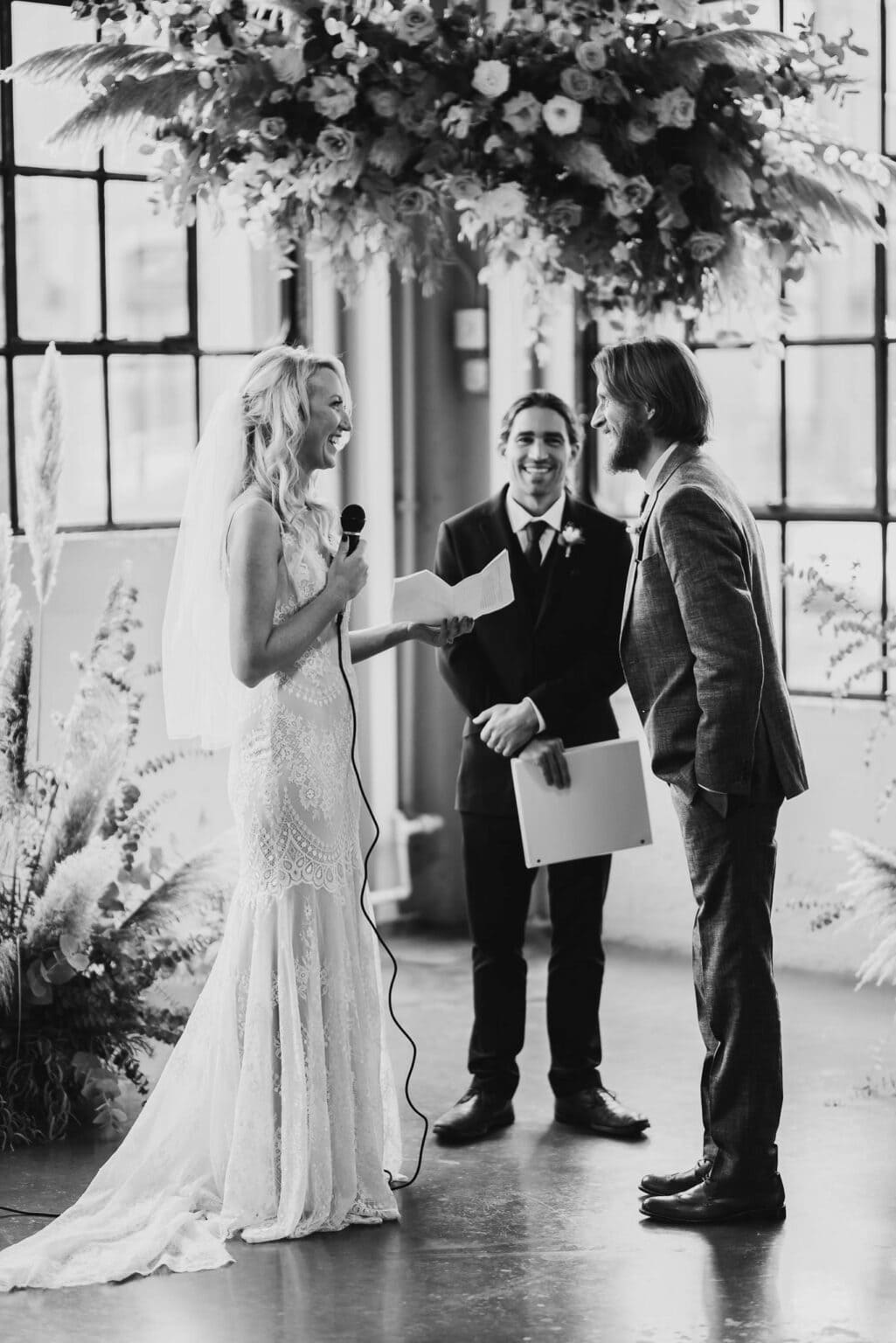 Bride and groom smiling during a wedding ceremony, with officiant in the background under a floral arrangement at Castaway Portland. Black and white.