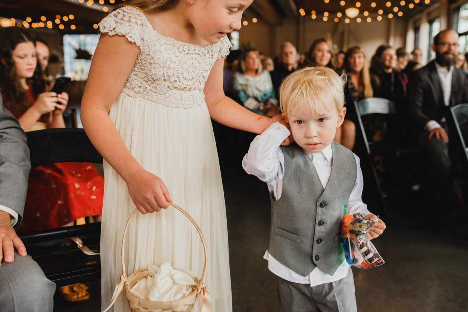 A young girl in a white dress, holding a basket, walks with a small boy in a gray suit at an indoor event. It seems like they might be capturing cherished wedding photos at Castaway Portland.