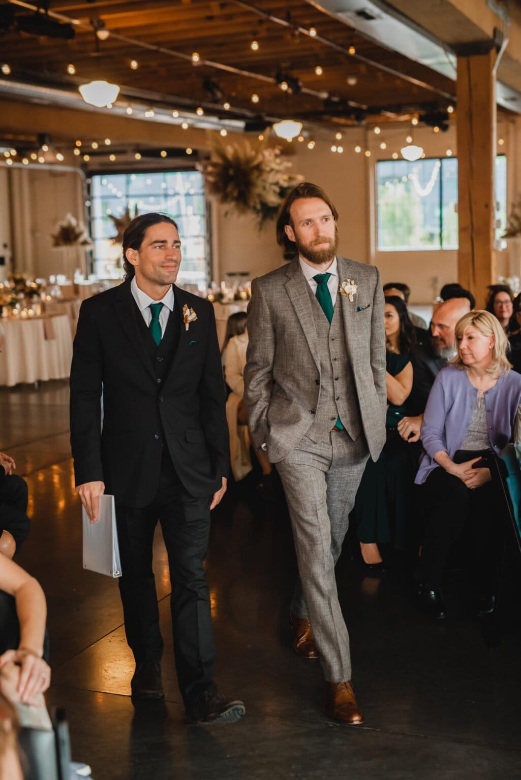 Two men in suits walk down an aisle at a wedding, while seated guests watch in a warmly lit venue, capturing perfect moments for wedding photos at Castaway Portland.