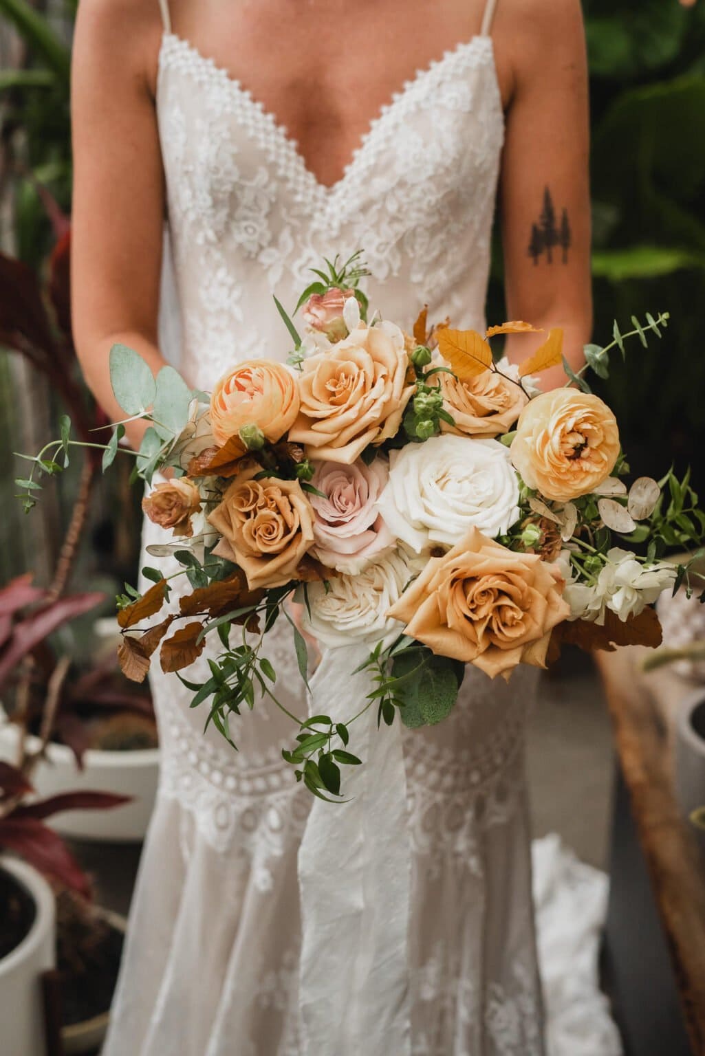 The bride, elegantly dressed in lace, holds a bouquet of vibrant orange and white roses with lush greenery. She gracefully poses for portraits at Pomerius Nursery before a wedding at the enchanting Castaway Portland.