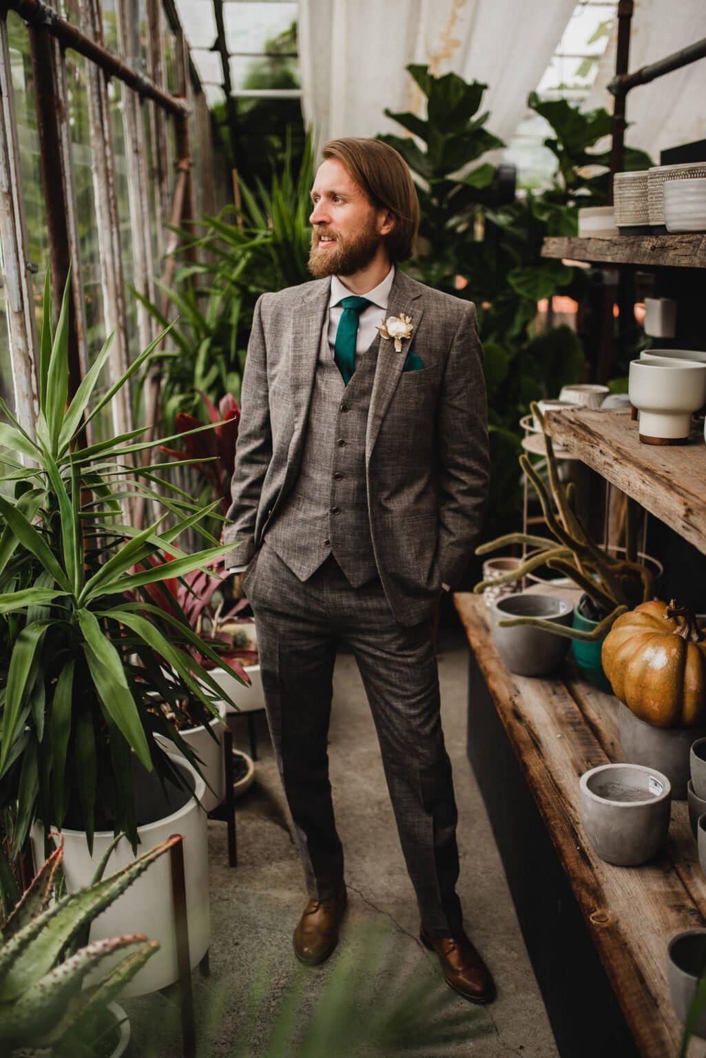 A man in a gray suit stands in a greenhouse at Pomerius Nursery, surrounded by plants and pots, capturing portraits before a wedding at Castaway Portland.