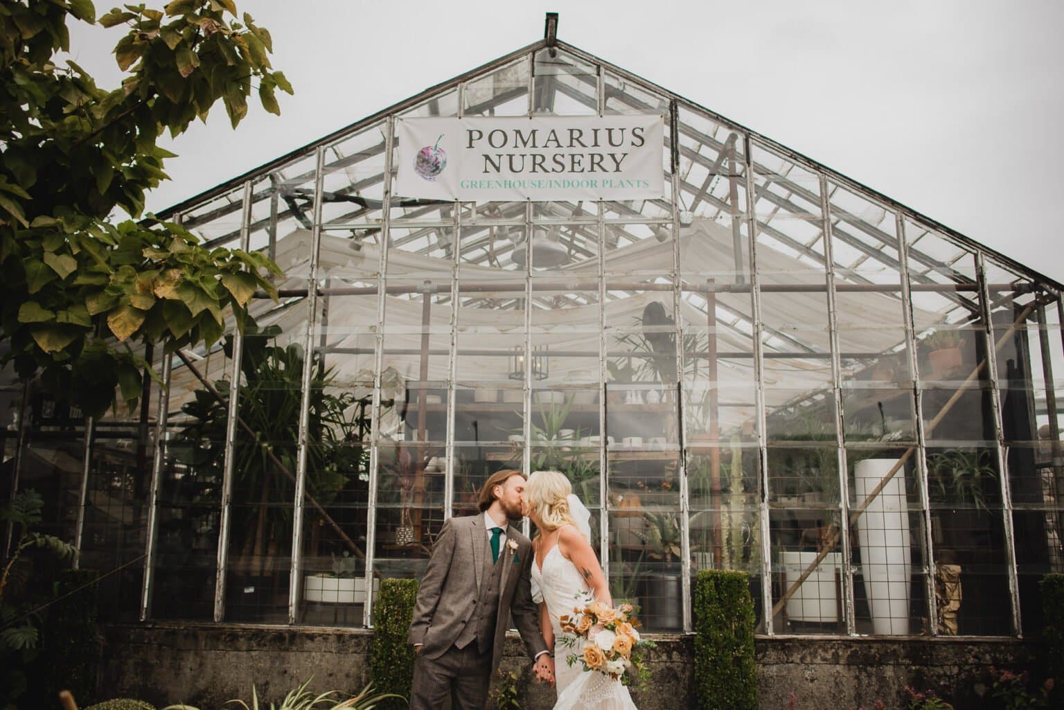 A couple shares a kiss in front of a greenhouse with a Pomarius Nursery sign, surrounded by lush greenery, capturing portraits at Pomarius Nursery before their wedding at Castaway Portland.