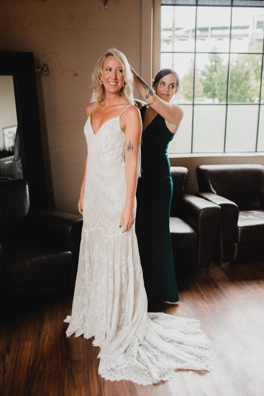 The bride in a lace dress gets her hair adjusted by another woman in a dimly lit room with sofas and a window, capturing the moment for wedding photos at Castaway Portland.