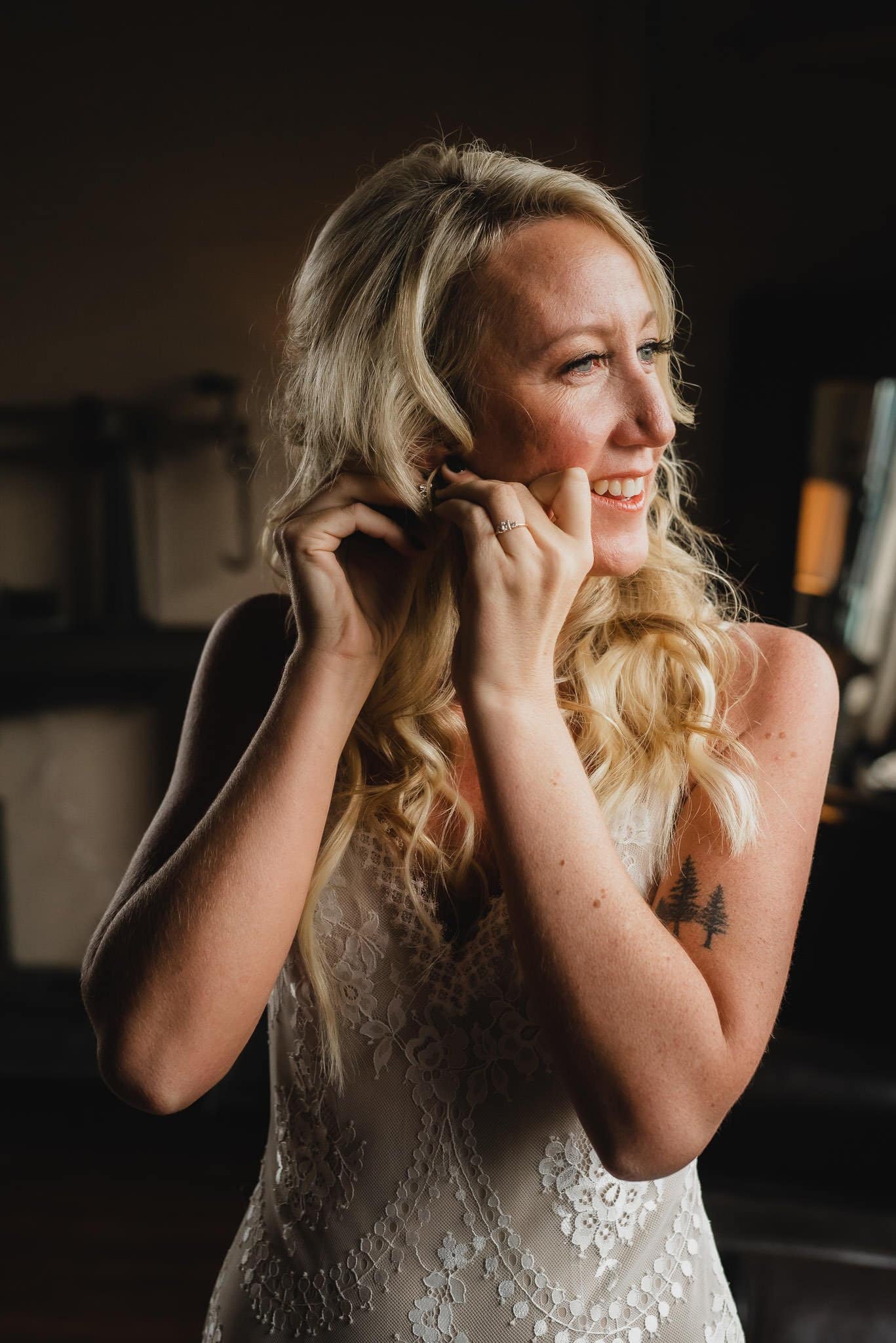 The bride in a white lace dress smiles as she adjusts her earring, with her side tattoo visible, ready for wedding photos at Castaway Portland.
