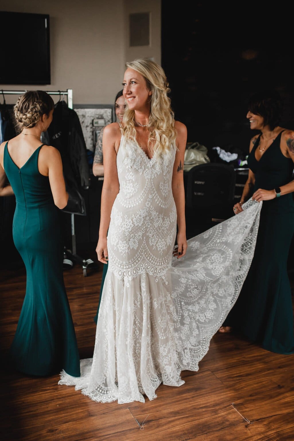 The bride, dressed in a lace gown, smiles as bridesmaids in green dresses assist with her train. Capturing the joy of this moment are the wedding photos at Castaway Portland.